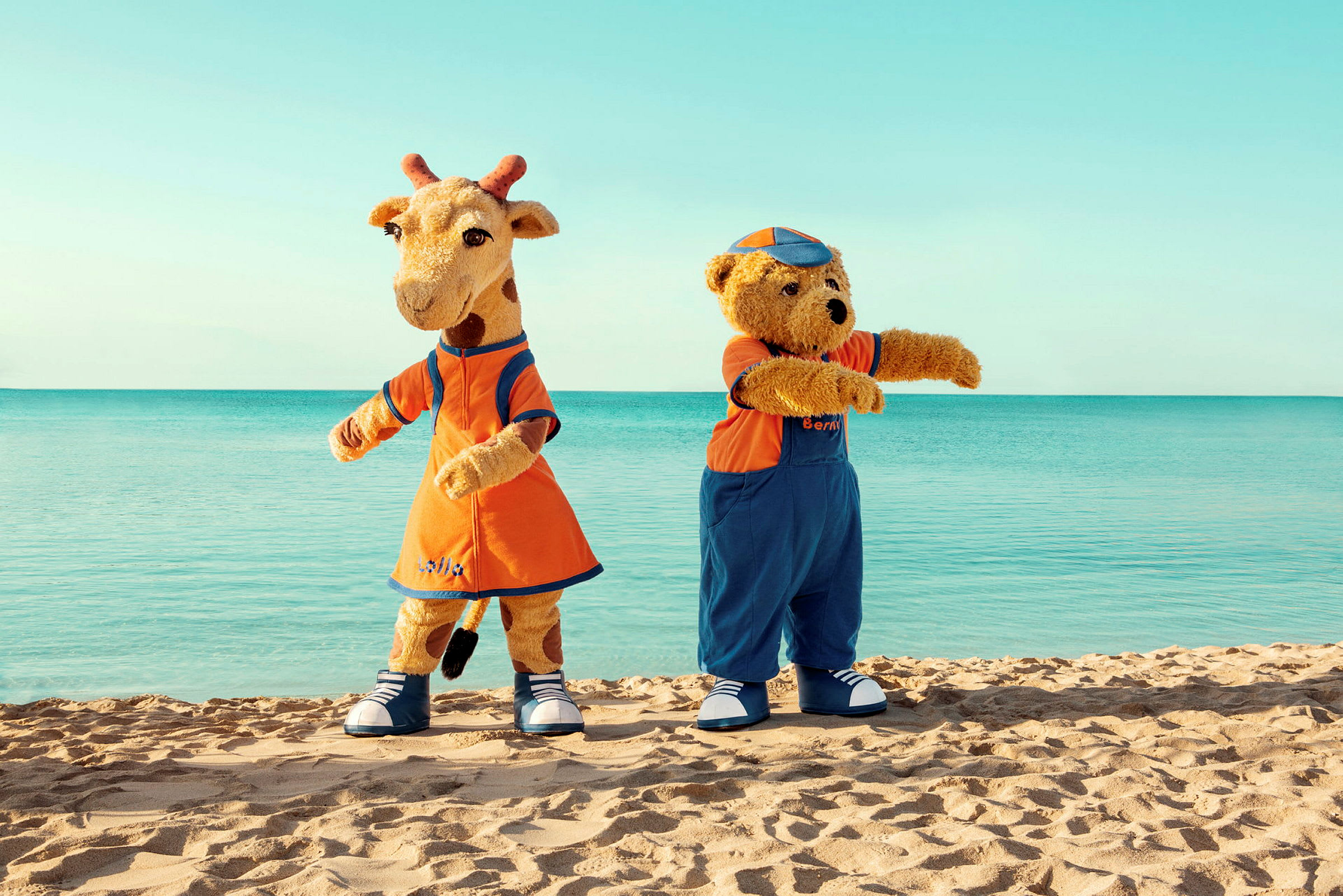 To maskotter, en giraf og en bjørn, står på en strand med havet i baggrunden. (AI-genereret tekst)