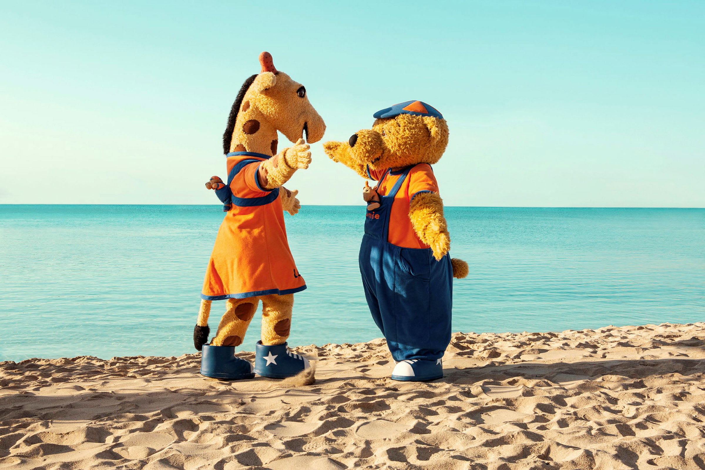 To maskotter, en giraf og en bjørn, står på en sandstrand med havet i baggrunden. (AI-genereret tekst)