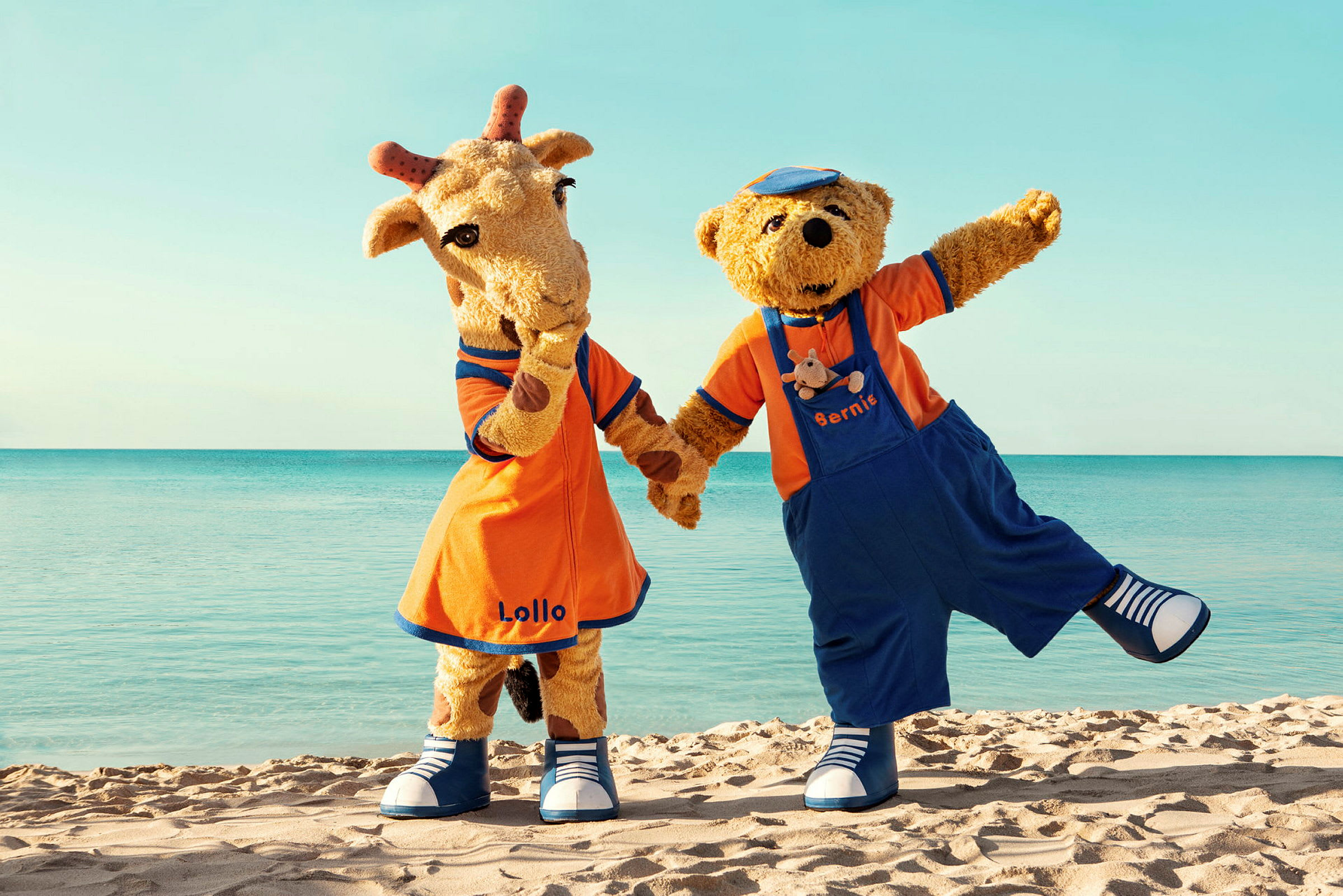 Maskotterne Lollo og Bernie står på en sandstrand foran havet. (AI-genereret tekst)