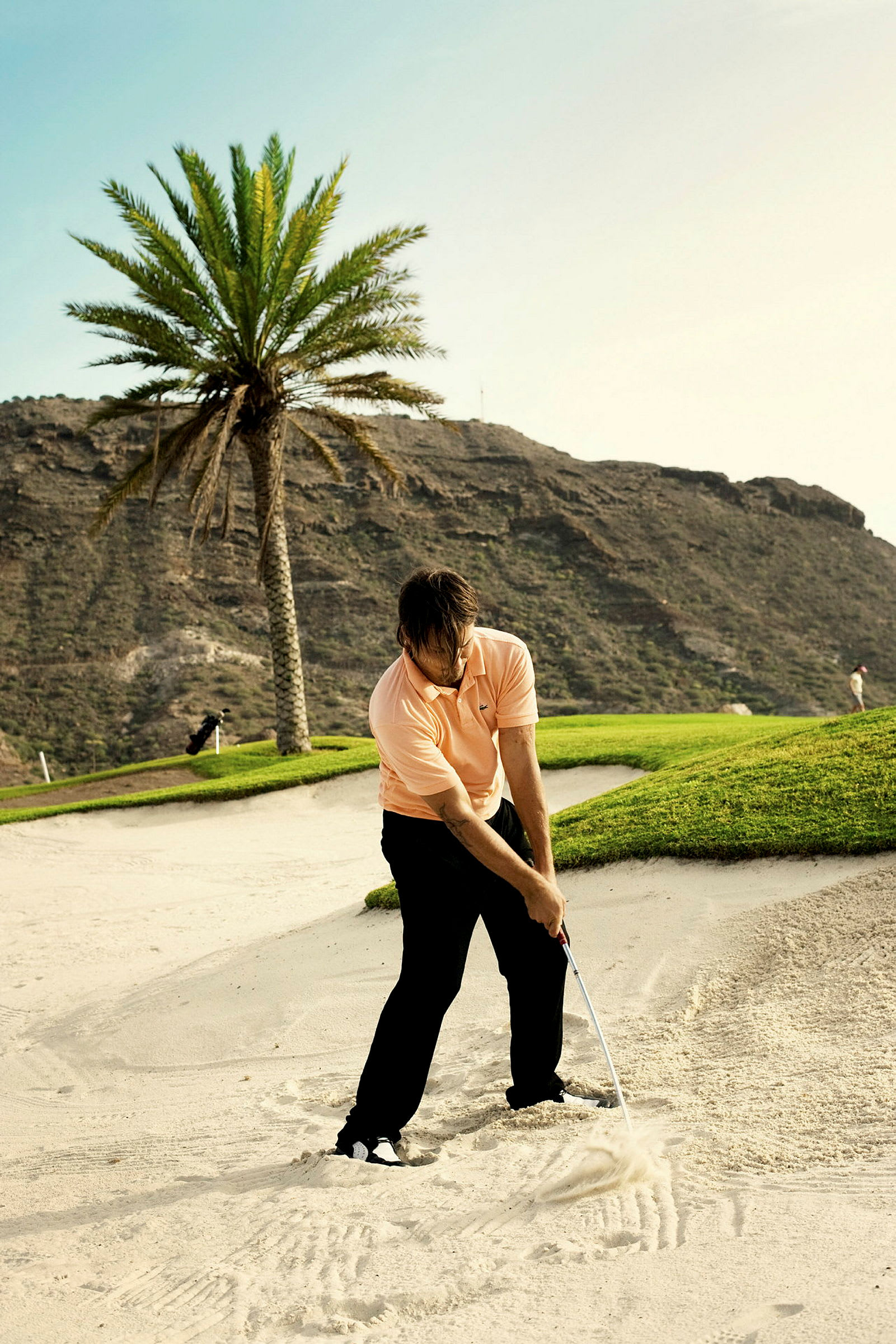 En person spiller golf i en sandbunker med en golfkølle. I baggrunden ses en palme og bjerge. (AI-genereret tekst)