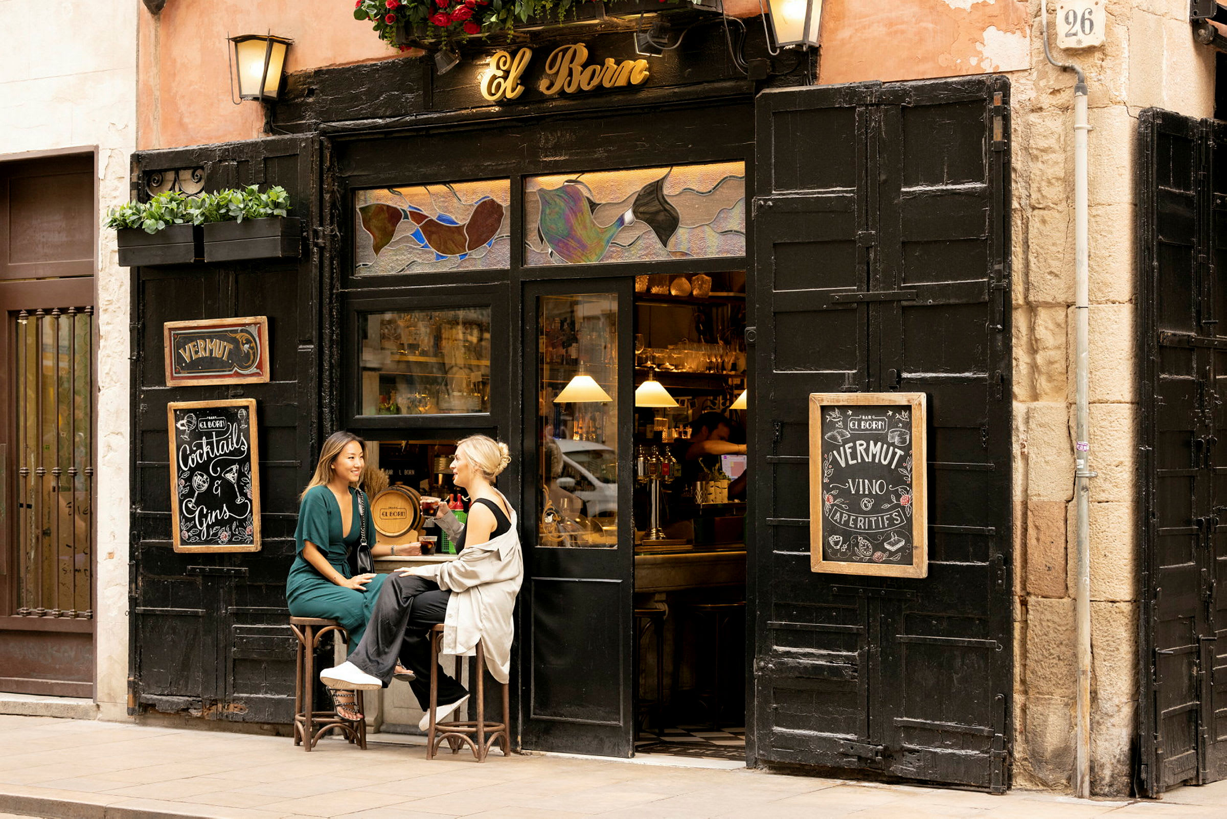 To personer sidder på skamler foran en lille bar med skiltet 'El Born'. (AI-genereret tekst)
