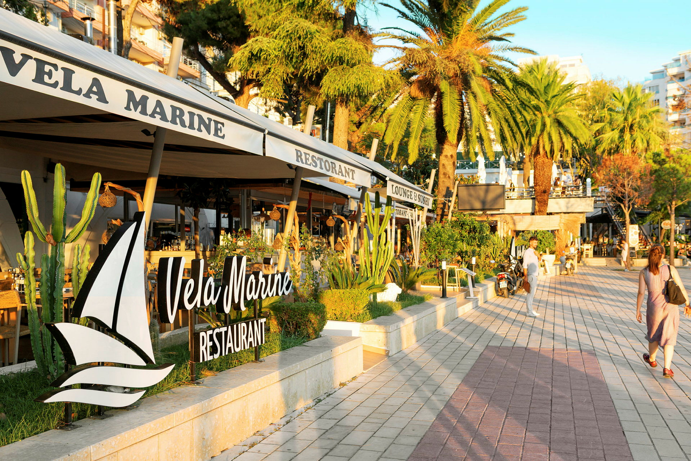 En strandpromenade med palmer og restauranter. Skiltet "Vela Marine Restaurant" er tydeligt. (AI-genereret tekst)