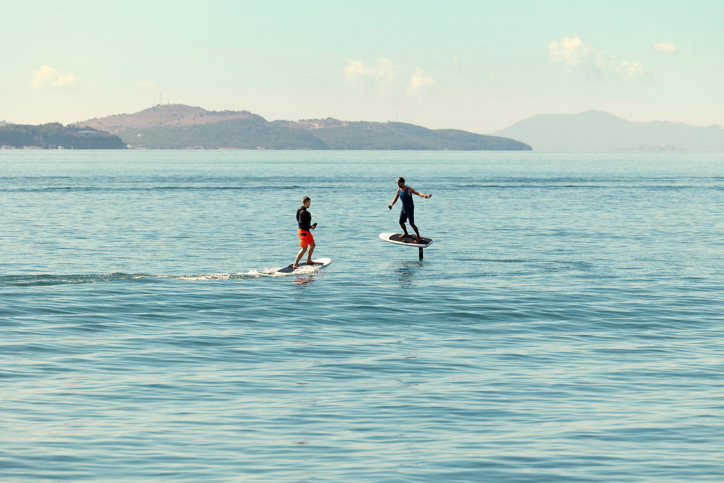 To personer står på elektriske surfbrætter på en rolig havoverflade med bjerge i baggrunden. (AI-genereret tekst)