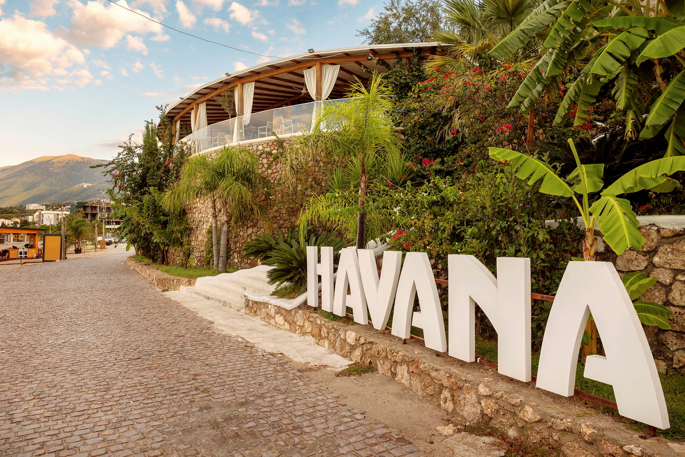 Skriven tekst med 'HAVANA' ved en tropisk bygning ved siden af brostensvej. (AI-genereret tekst)