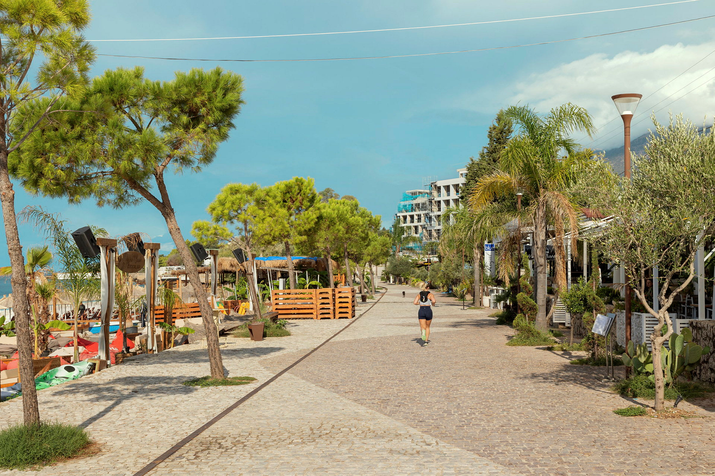 En brostensbelagt promenade omgivet af træer, restauranter og et strandområde. (AI-genereret tekst)