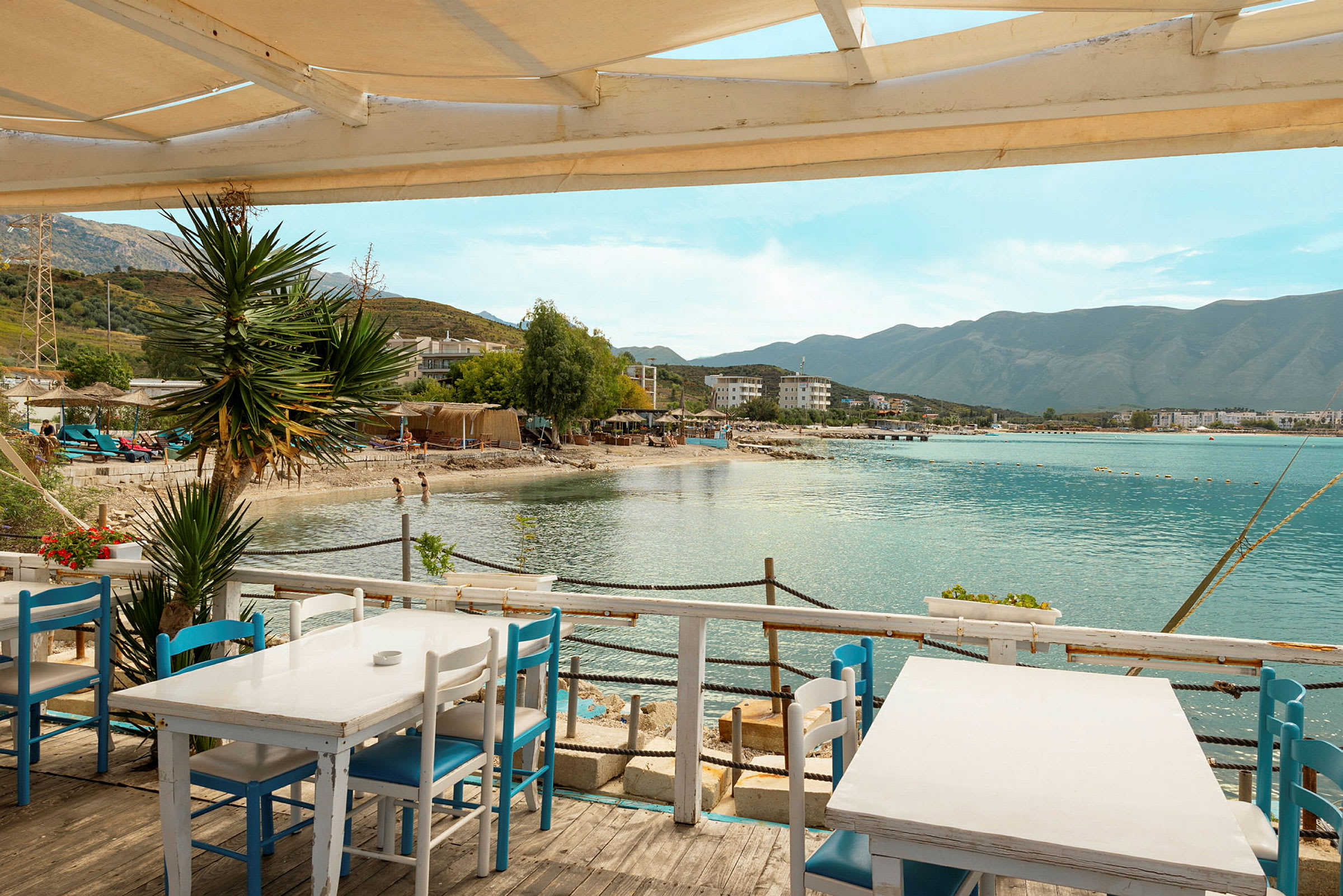 Udsigt fra en terrasse med hvide borde og blå stole, mod en strand, havet og bjerge i baggrunden. (AI-genereret tekst)