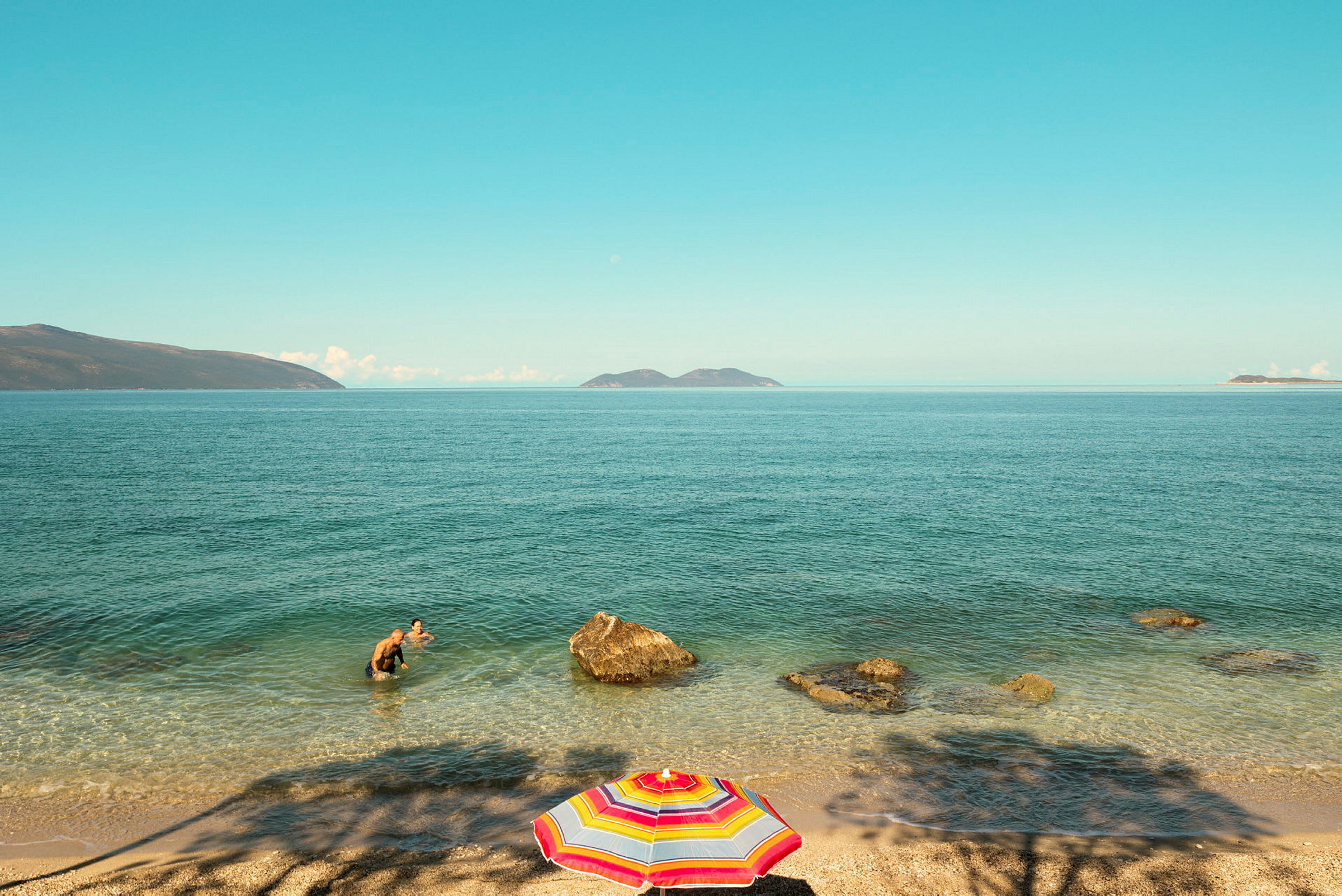 Stranden med en flerfarvet parasol, to personer bader i havet nær klipper. (AI-genereret tekst)