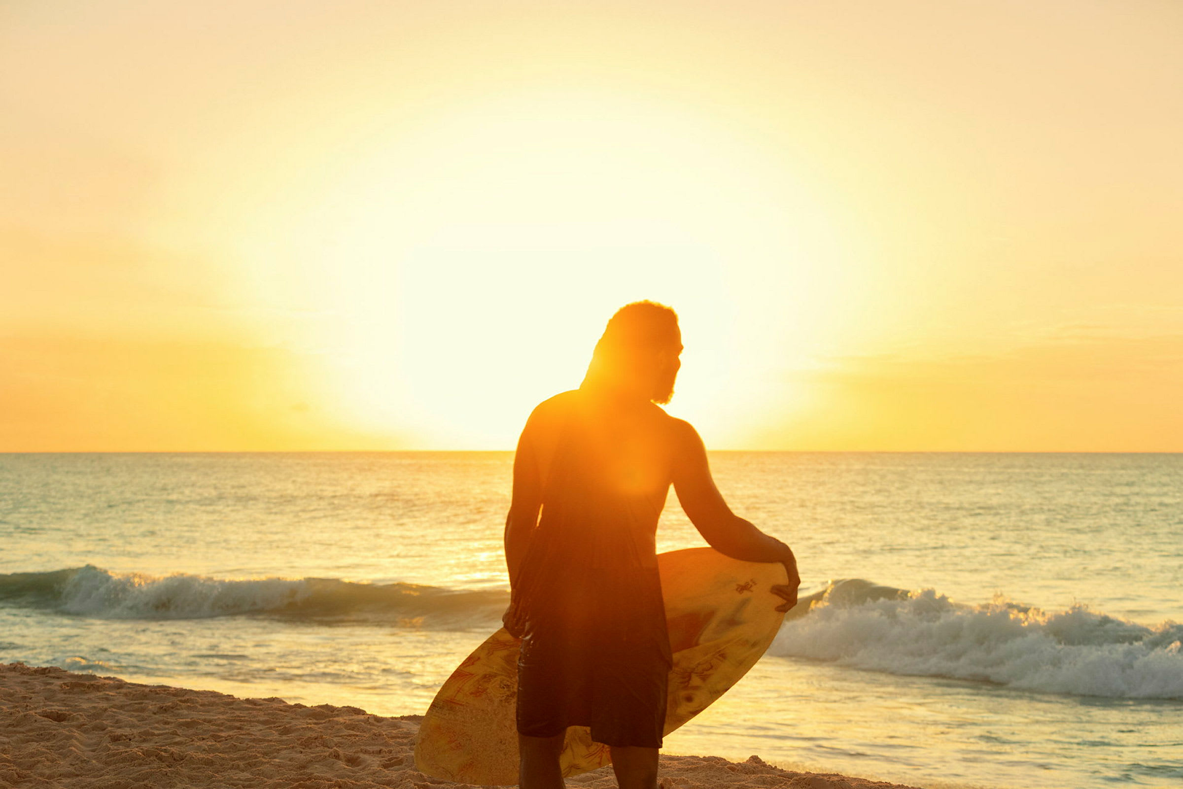 En person står på stranden ved havet, holder et surfbræt, og solen skinner klart i baggrunden. (AI-genereret tekst)