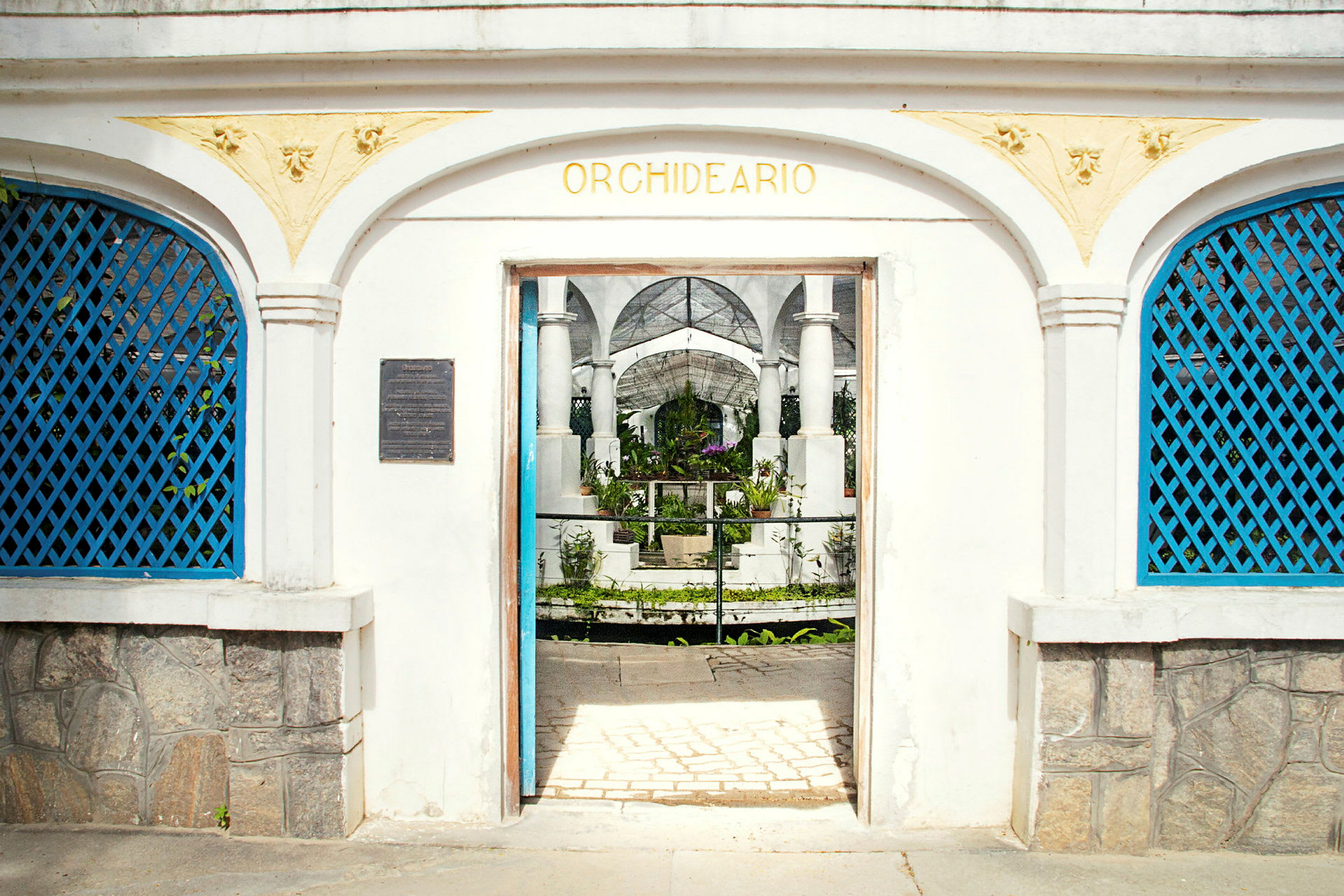 En hvid bygning med en veranda med buer og teksten "Orchideario" over indgangen, der fører ind til en have. (AI-genereret tekst)