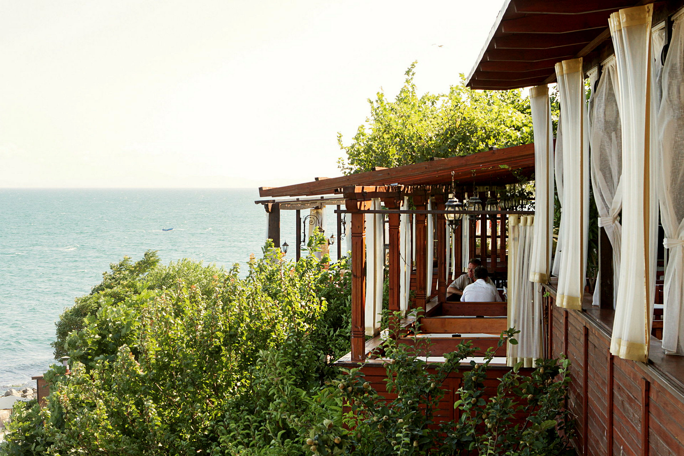 En restaurant med træpergola og hvide gardiner, omgivet af grønne områder, tæt på havet. (AI-genereret tekst)