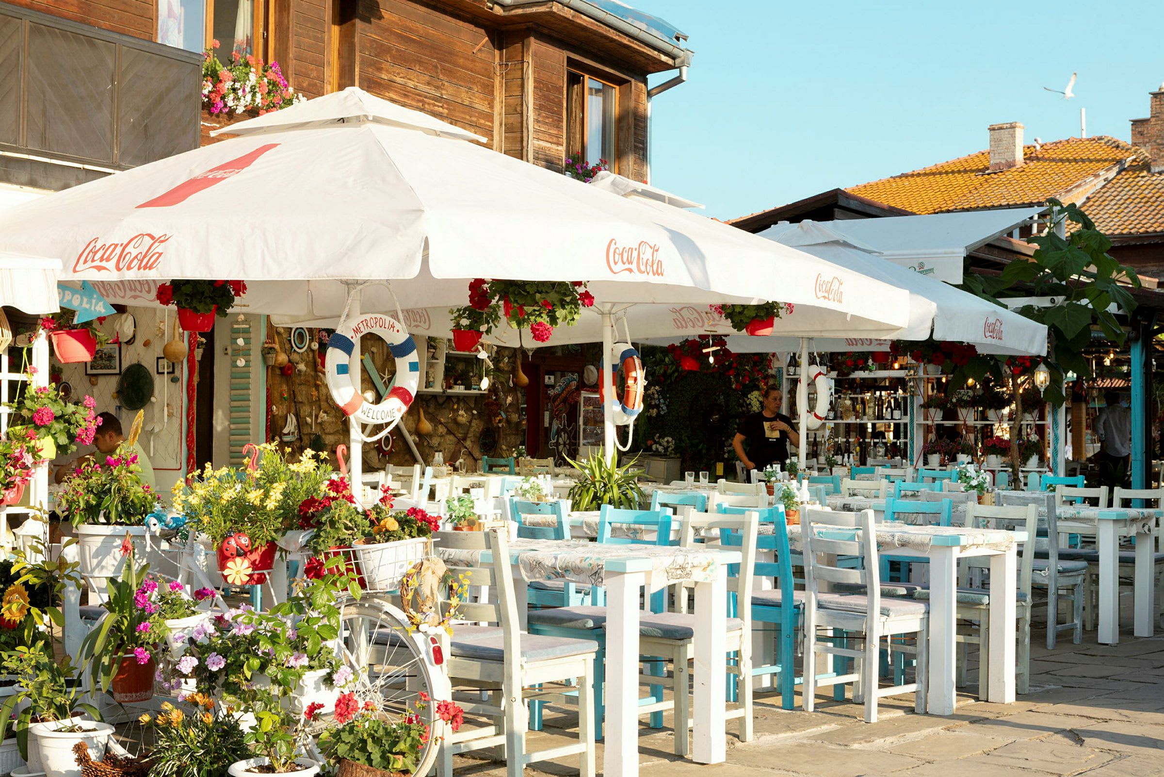 Billedet viser en udendørs café med blomster og hvide parasoller. (AI-genereret tekst)