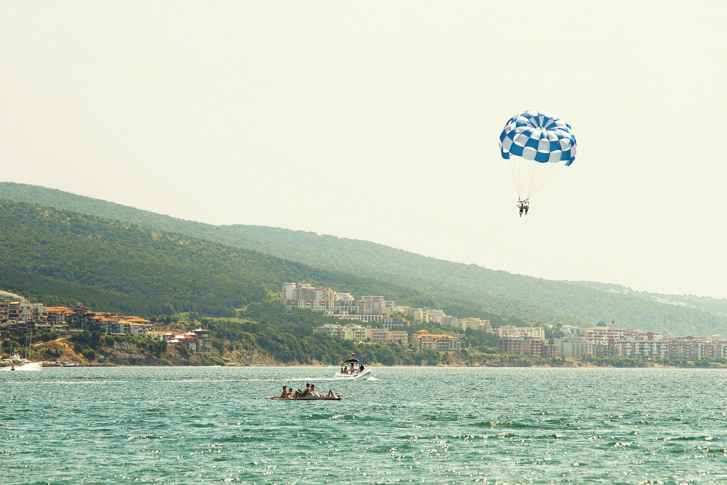 Personer der parasailer med en blå-hvid faldskærm over vandet ved en kyst. (AI-genereret tekst)