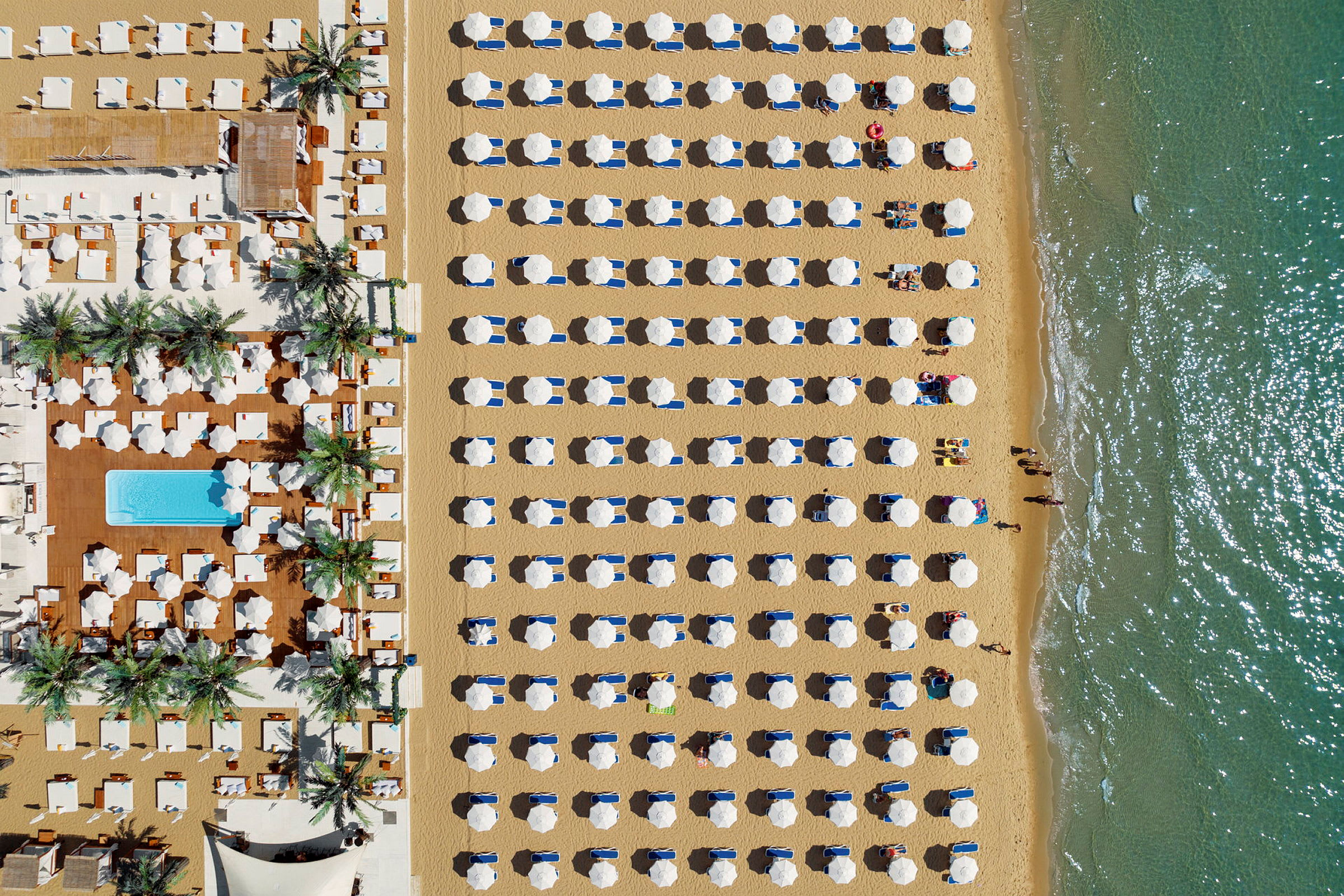 Luftfoto af en strand med arrangerede parasoller og solvogne tæt på vandet. (AI-genereret tekst)
