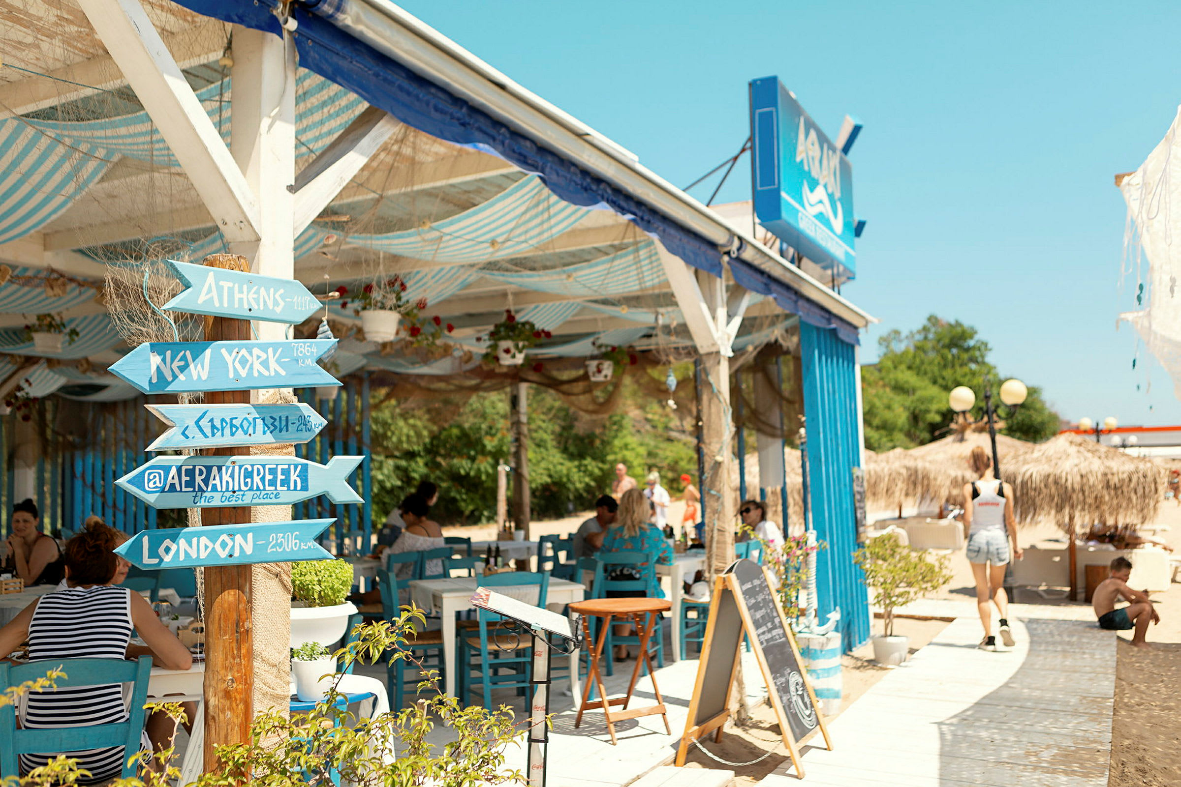 En strandnær restaurant med blå møbler og et træskilt med destinationsnavne. (AI-genereret tekst)