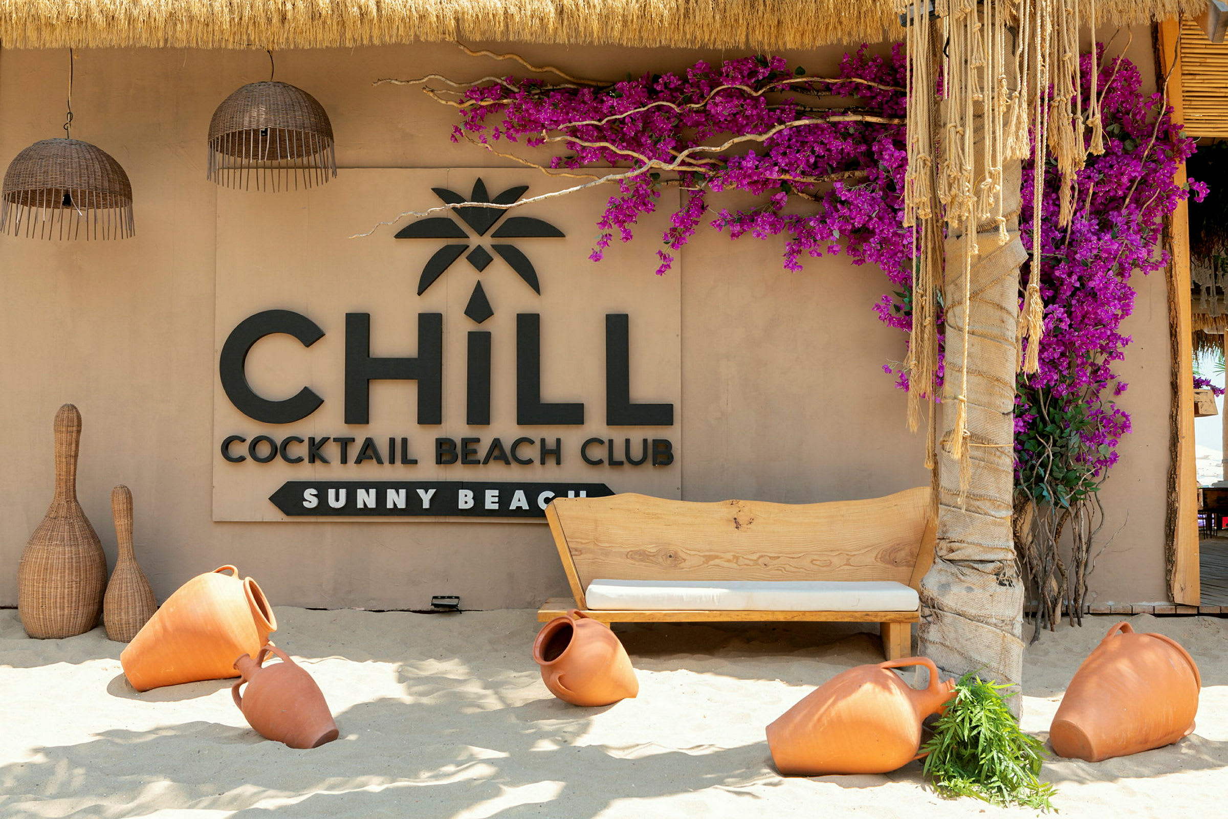 En strandbar med et skilt, der siger 'Chill Cocktail Beach Club Sunny Beach', omgivet af terrakrukker og farverige blomster. (AI-genereret tekst)