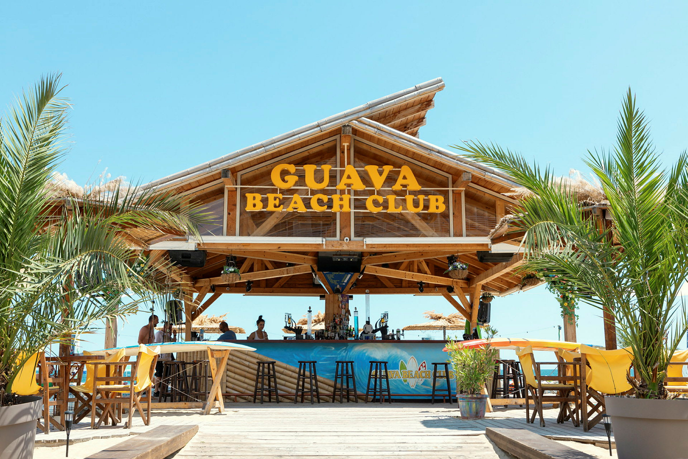 Billede af Guava Beach Club, en træbar med siddepladser, palmer og en klar himmel. (AI-genereret tekst)
