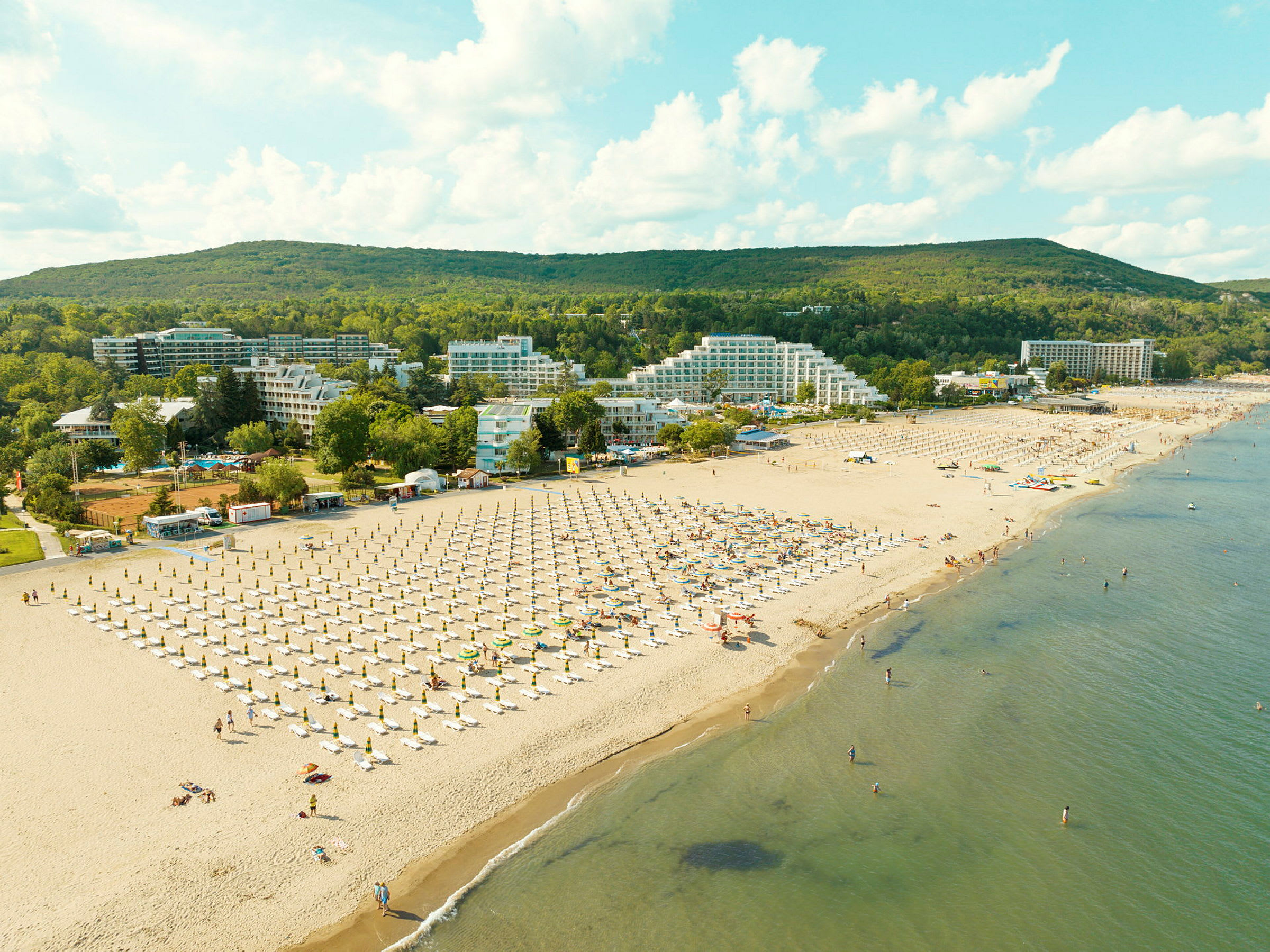 Strand med liggestole og parasoller, omgivet af hotelbygninger og grønne områder. (AI-genereret tekst)