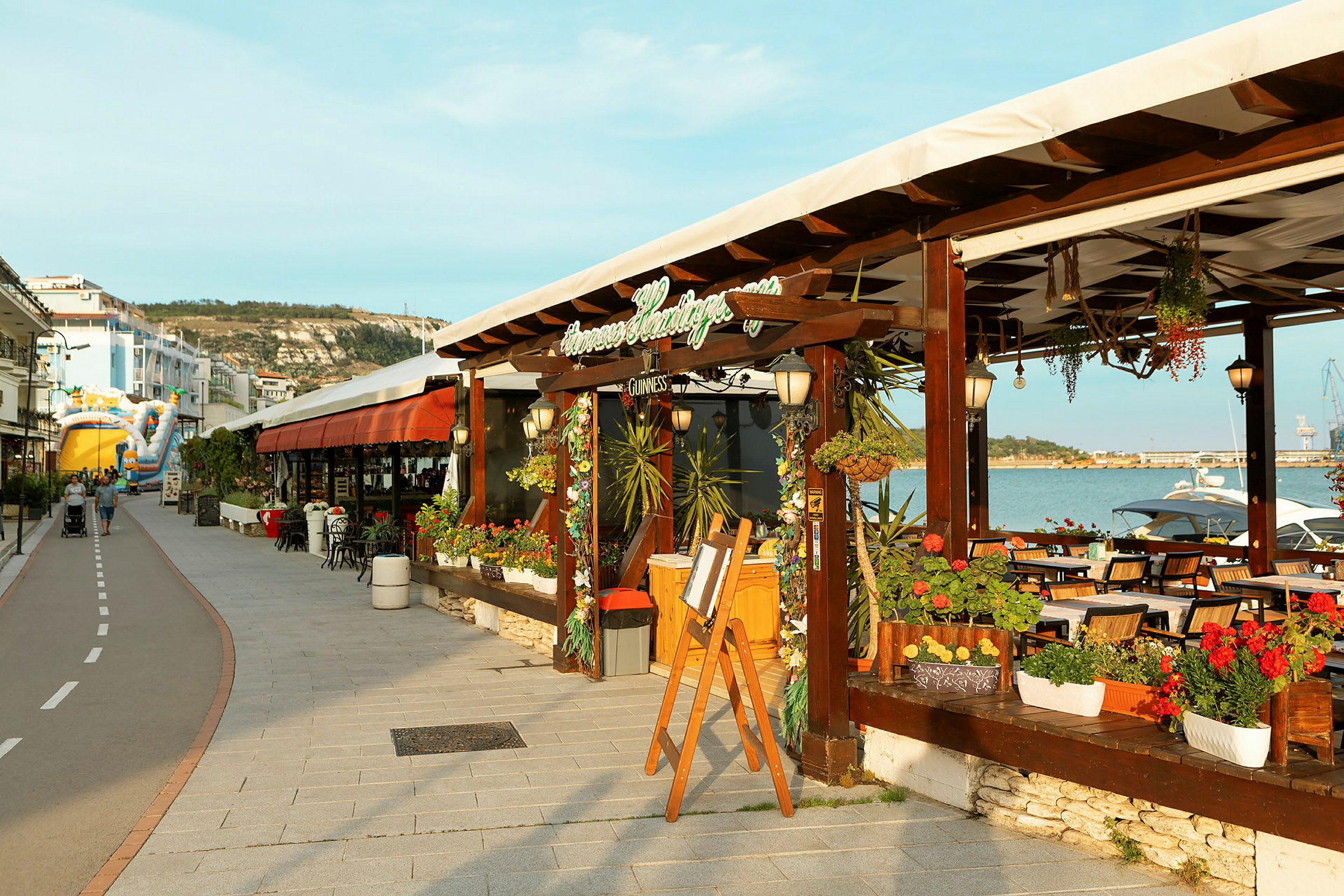 En udendørs café ved en strandpromenade med blomsterpotter og siddepladser. (AI-genereret tekst)