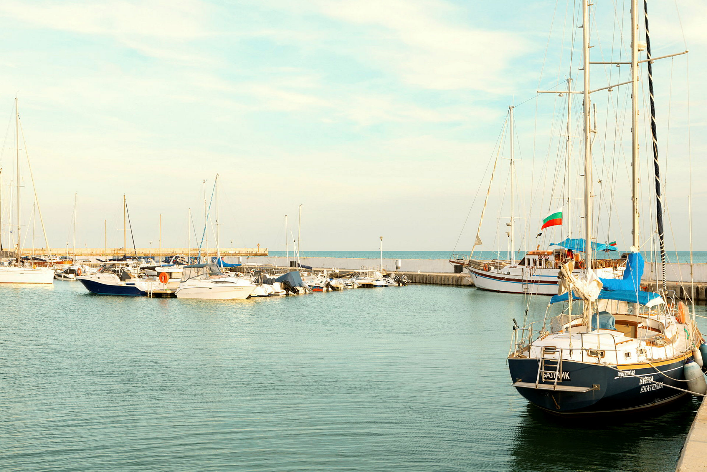 Små både og sejlskibe fortøjet i havnen med havet i baggrunden. (AI-genereret tekst)