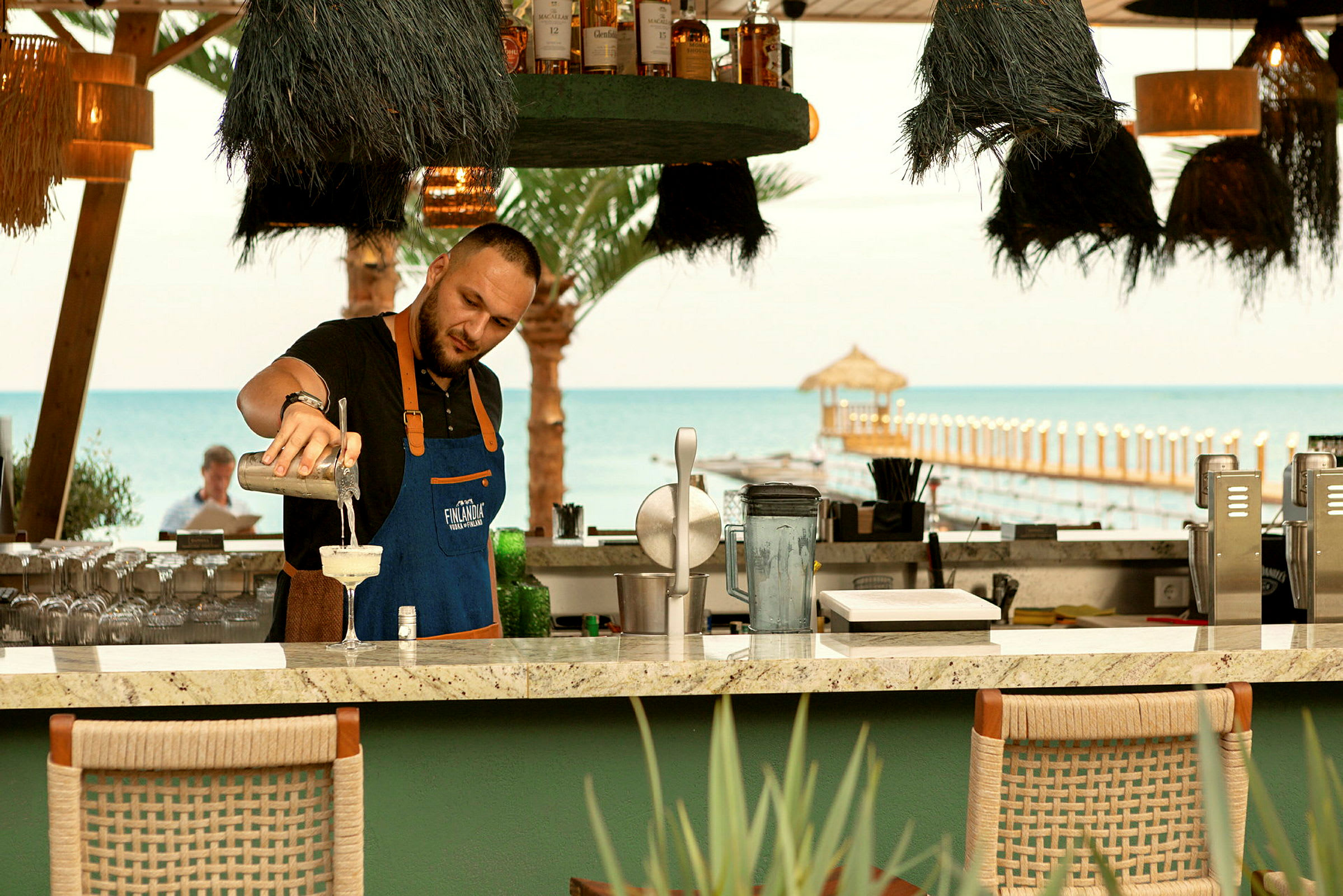En bartender ved en strandbar hælder en drink op med havet og en bro i baggrunden. (AI-genereret tekst)