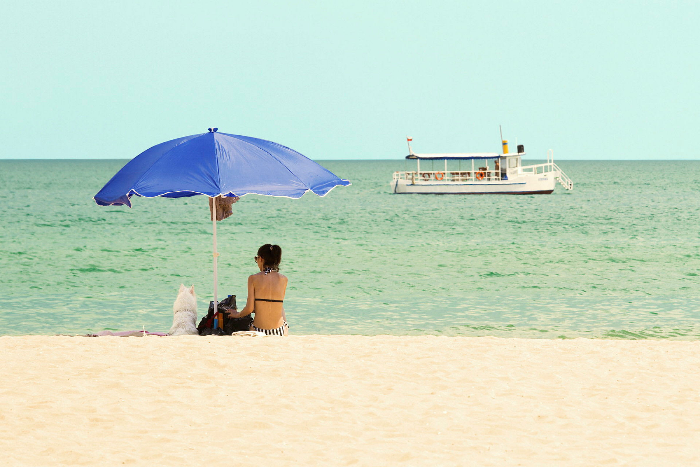 En person sidder under en blå parasol på en sandstrand med havet i baggrunden. En hvid båd ses på vandet. (AI-genereret tekst)