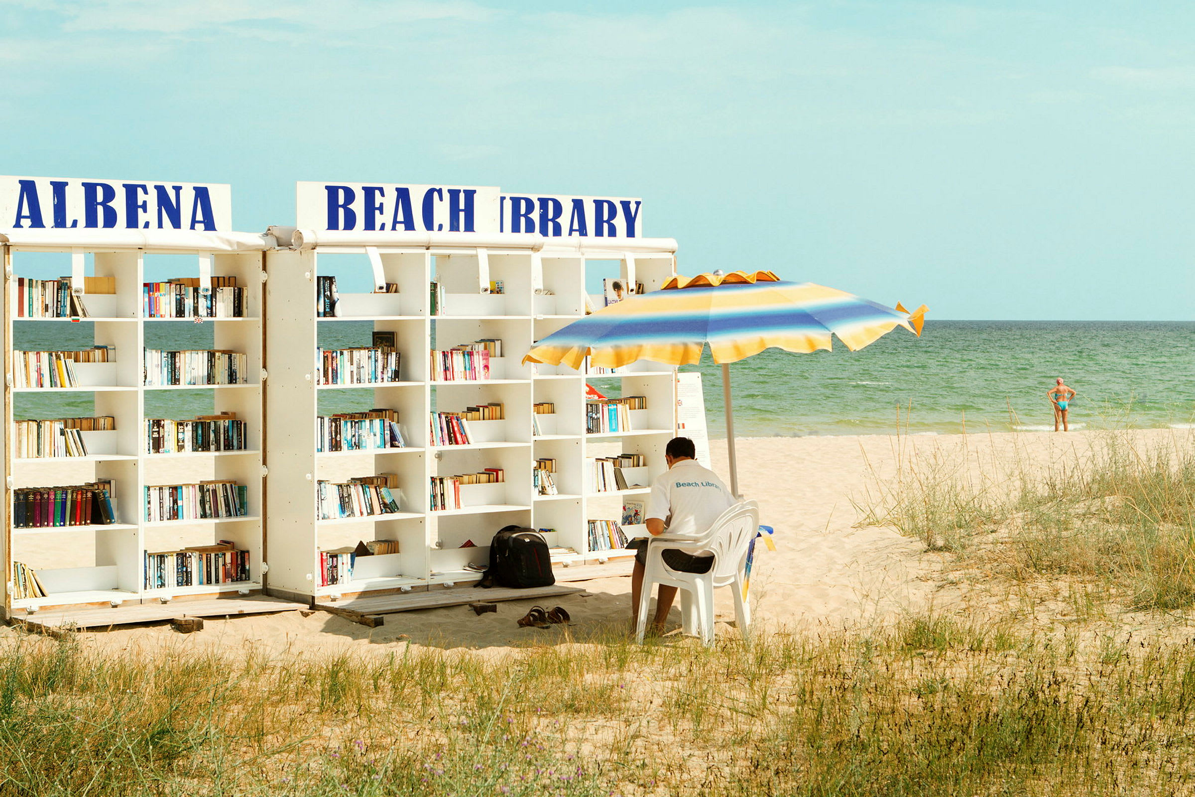 En strand med et udendørs bibliotek og en mand, der sidder under en parasol. (AI-genereret tekst)