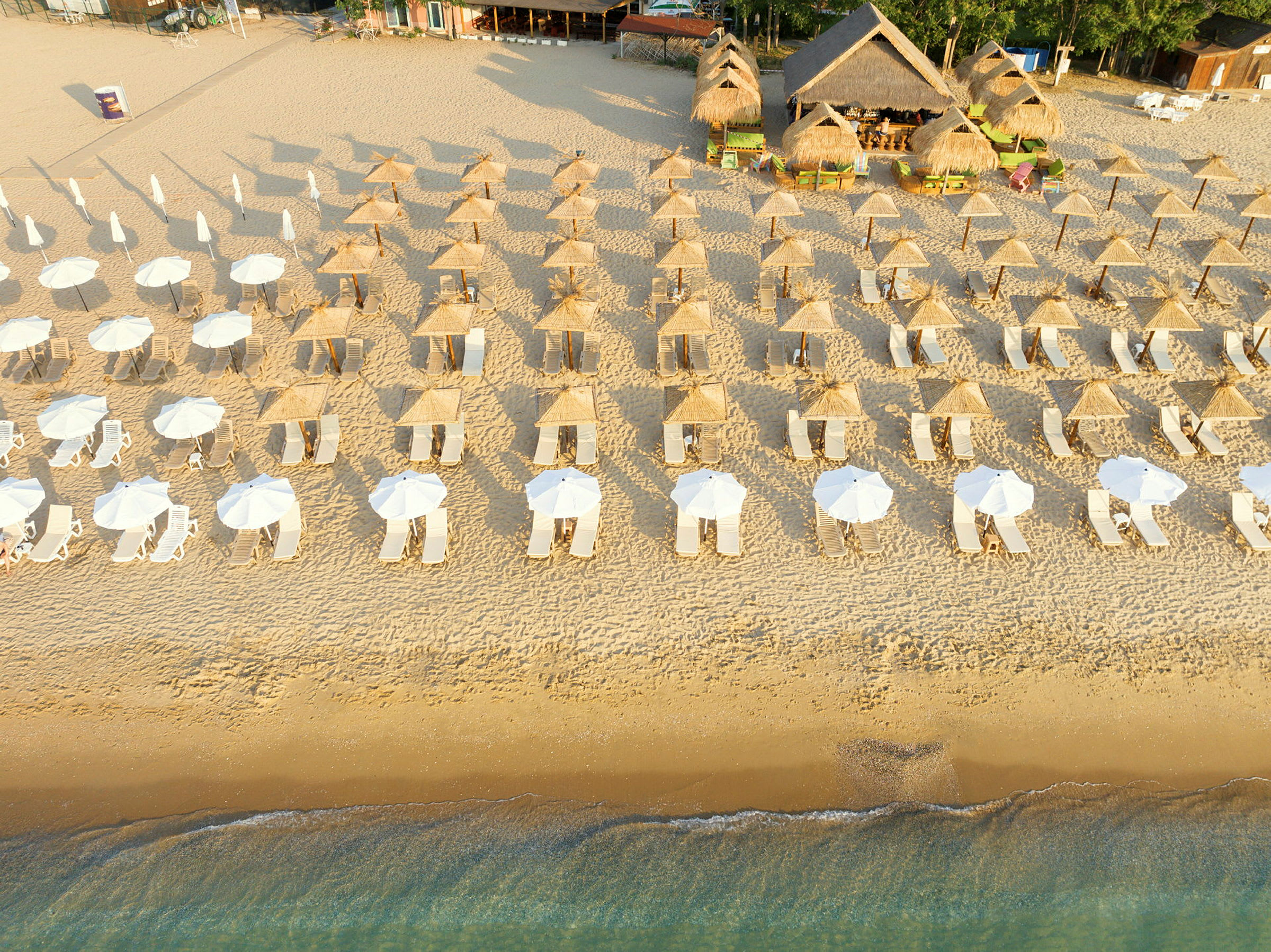 Luftfoto af en strand med rækker af liggestole og parasoller samt strandbar med stråtag i baggrunden. (AI-genereret tekst)