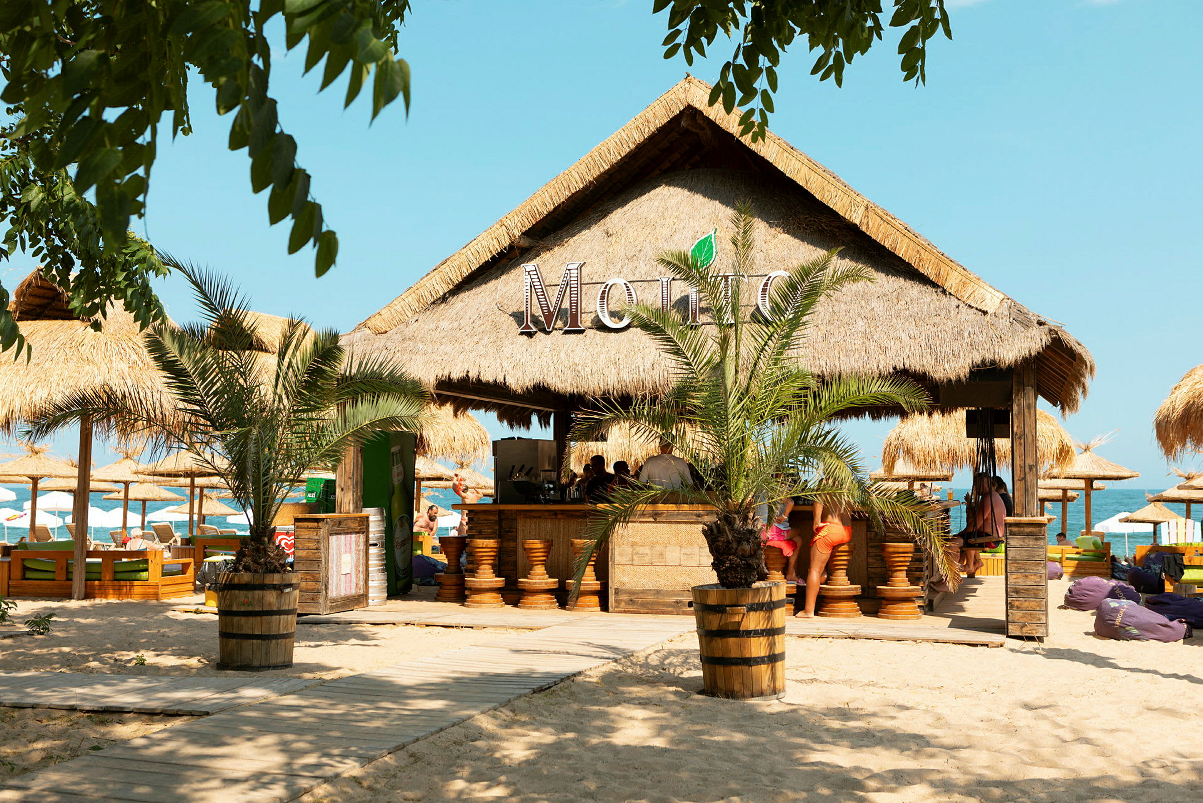 Strandbar med palmebladtag omgivet af sand og parasoller. (AI-genereret tekst)