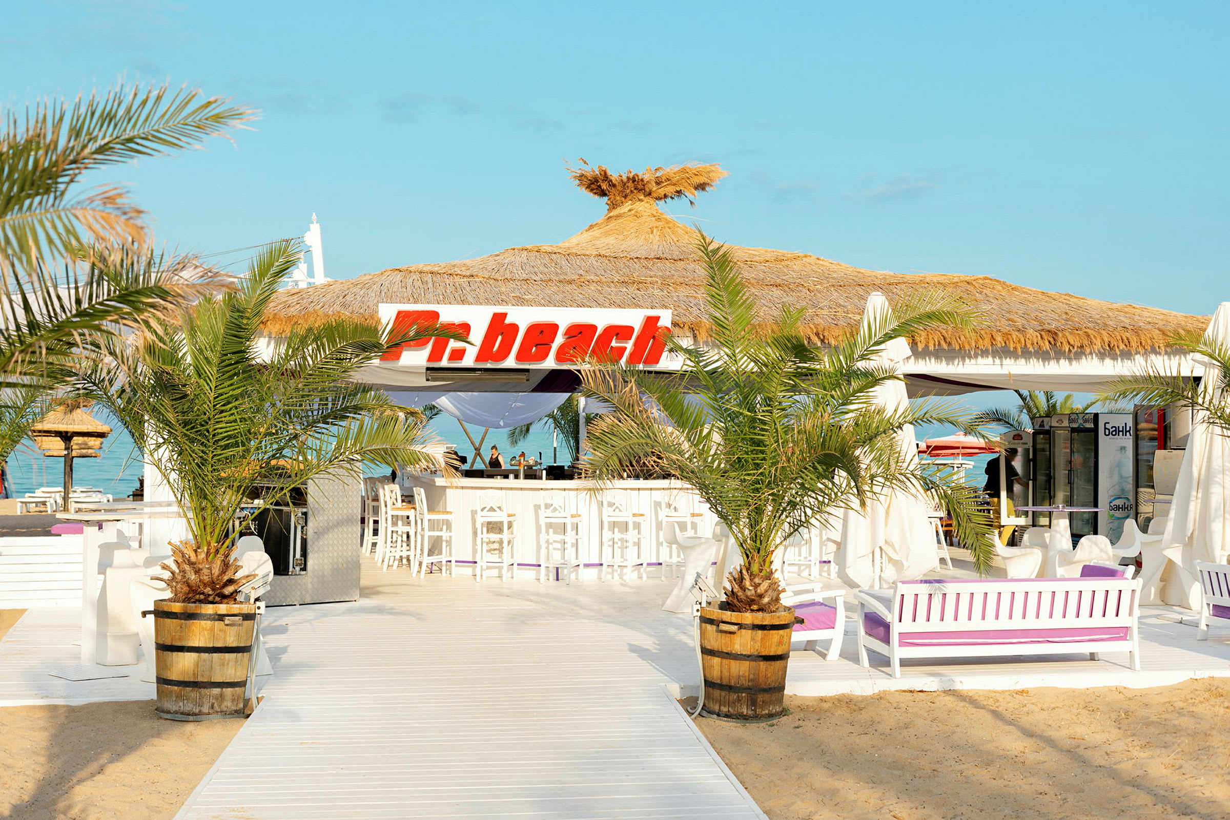 En strandbar med stråtækt tag og skilt med teksten 'Pn beach', omgivet af palmer og hvidt interiør. (AI-genereret tekst)