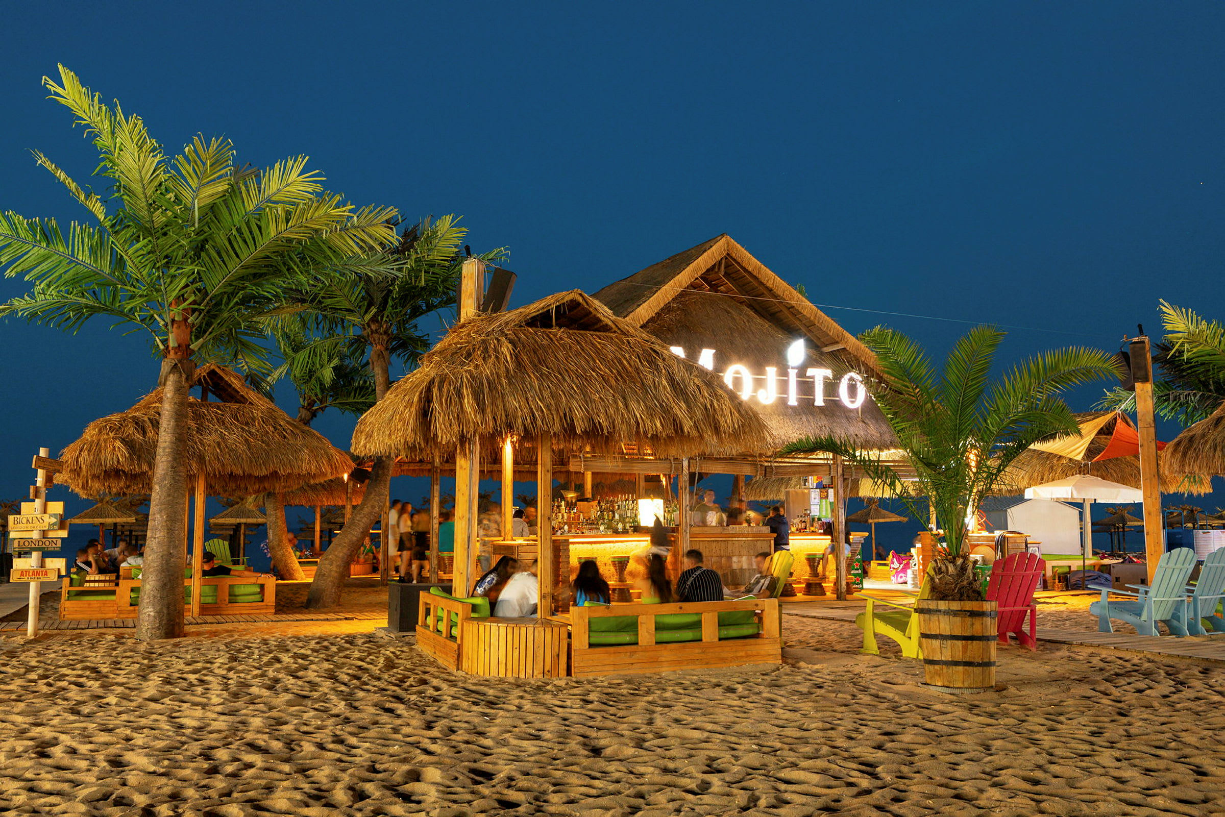 Strandbar med palmebladstag, stole og borde på sandet grund om aftenen. (AI-genereret tekst)