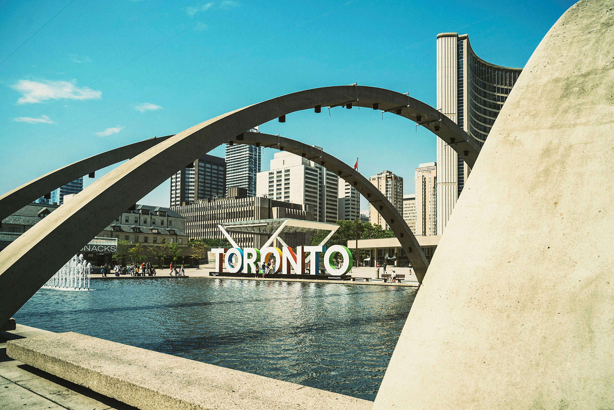 Et udsigt over Nathan Phillips Square i Toronto med en springvand, buer og byens skyline i baggrunden. (AI-genereret tekst)