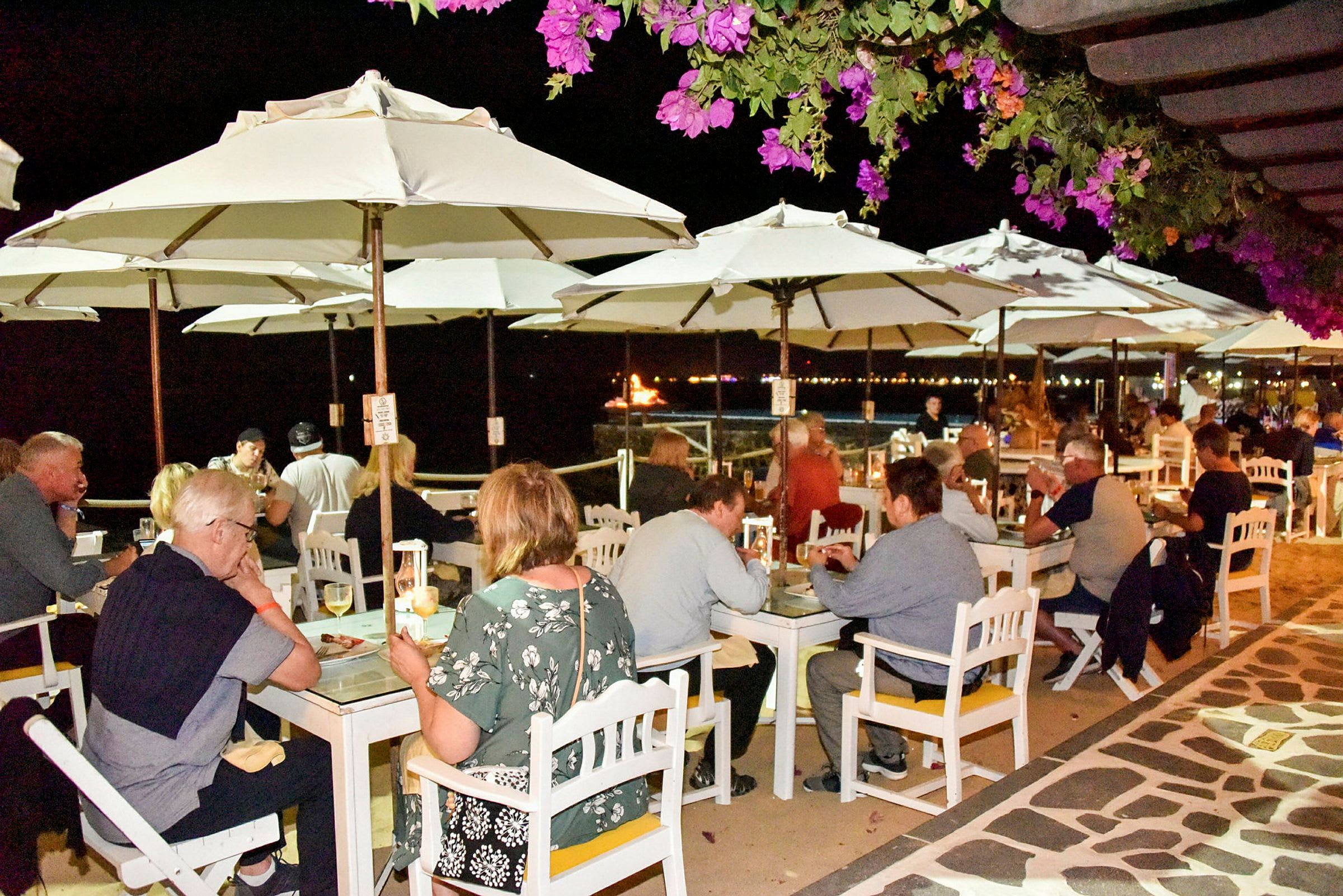 Mennesker sidder ved udendørsborde under hvide parasoller på en aftenrestaurant, med blomstrende planter i forgrunden og havet i baggrunden. (AI-genereret tekst)