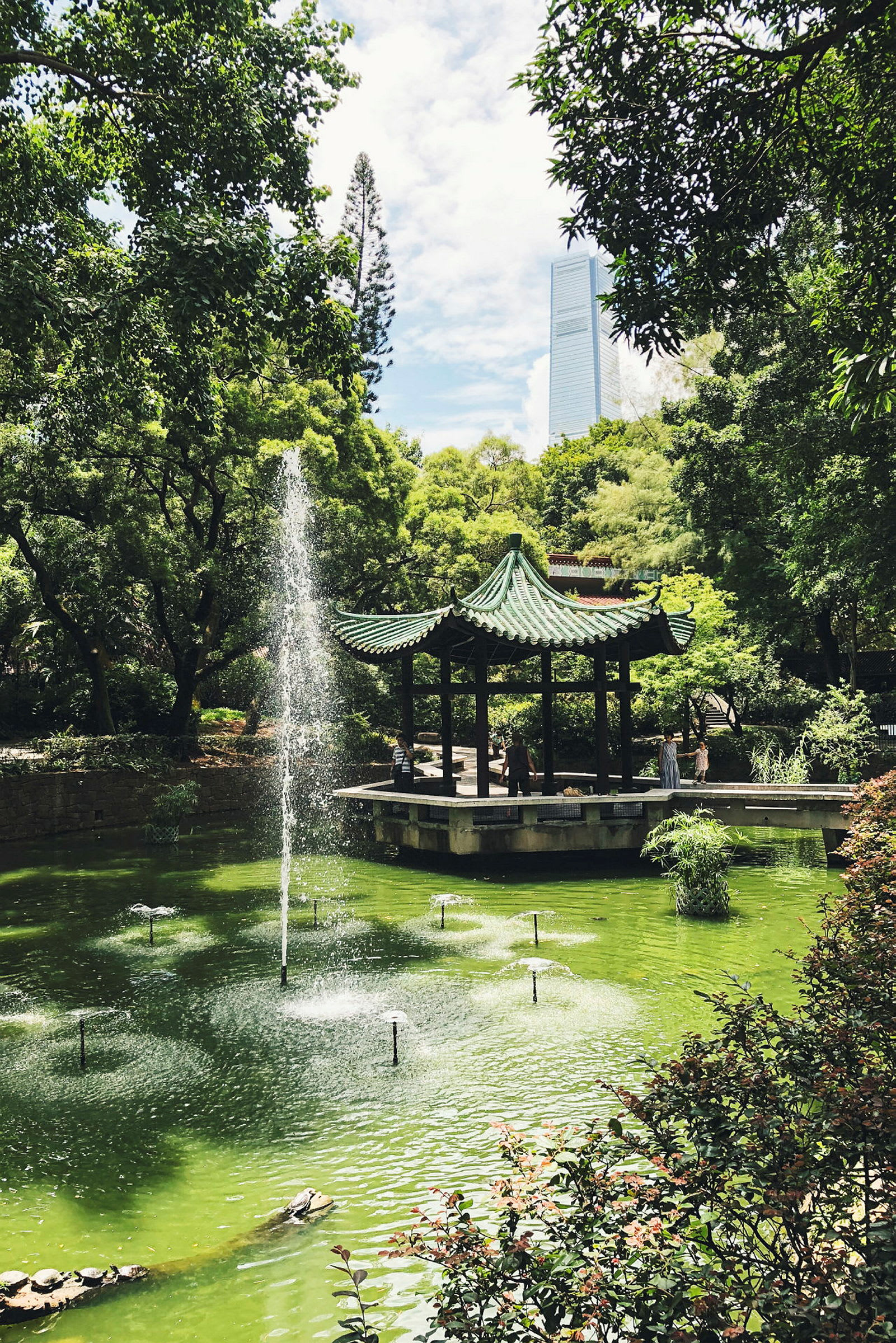 En asiatisk-inspireret pavillon ved en grøn dam omgivet af træer. (AI-genereret tekst)