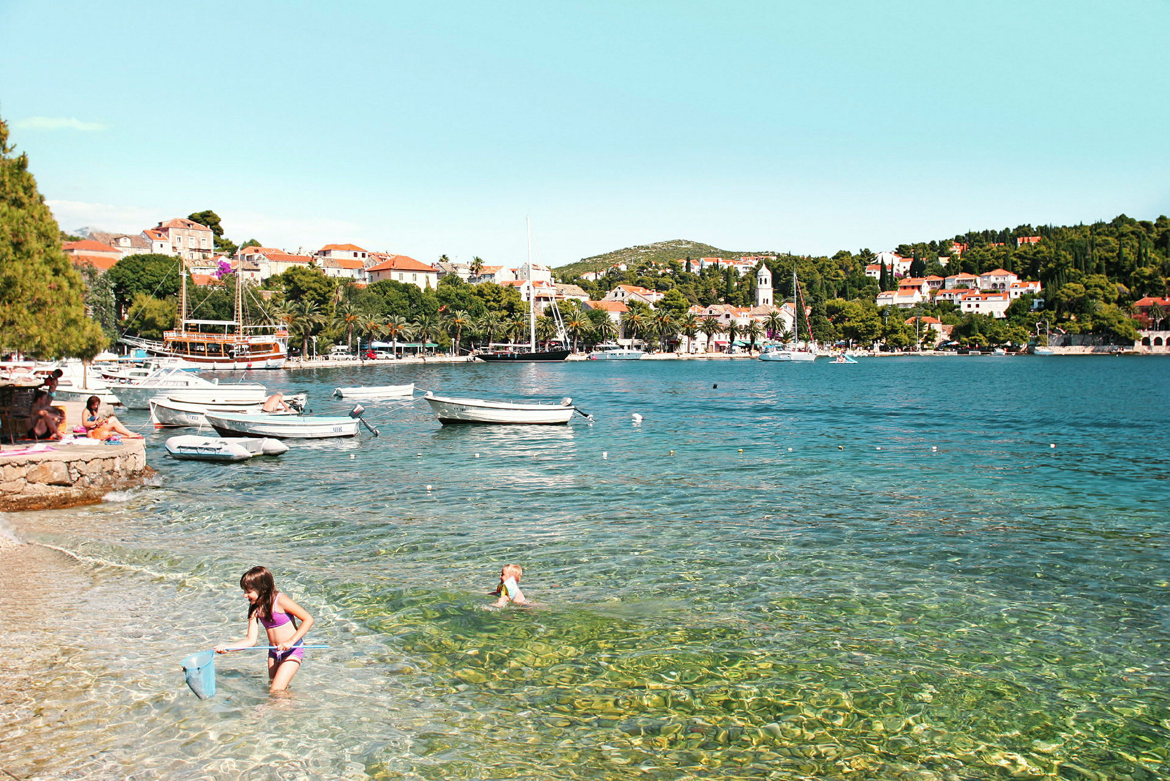 Strand med klart vand, småbåde og en by i baggrunden. (AI-genereret tekst)