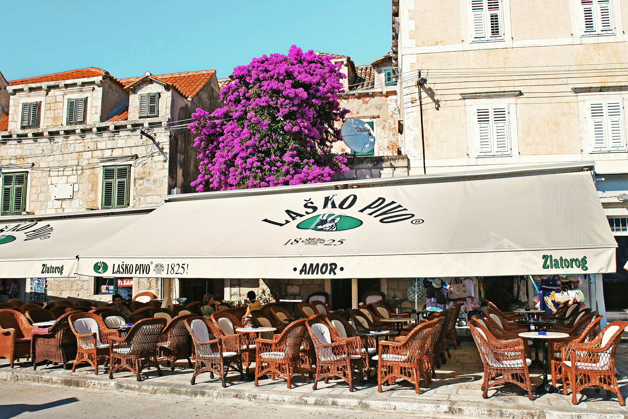 En udendørs café med kurvestole og borde under et stort tag, samt en bygning med vinduer og en blomstrende plante. (AI-genereret tekst)