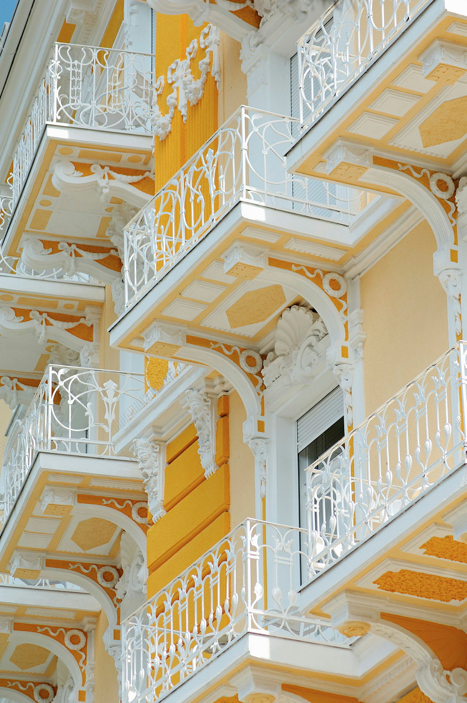 Detaljeret facade med gule vægge, hvide dekorationer og altaner med slyngende gelændere. (AI-genereret tekst)