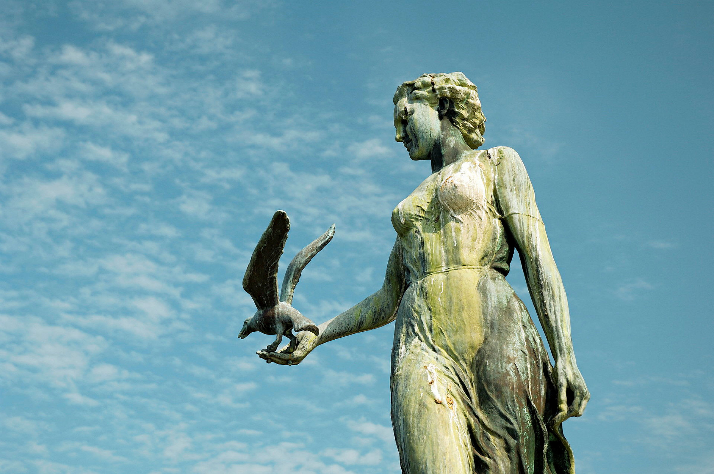 Statue af en kvinde med en fugl i hånden mod en blå himmel. (AI-genereret tekst)