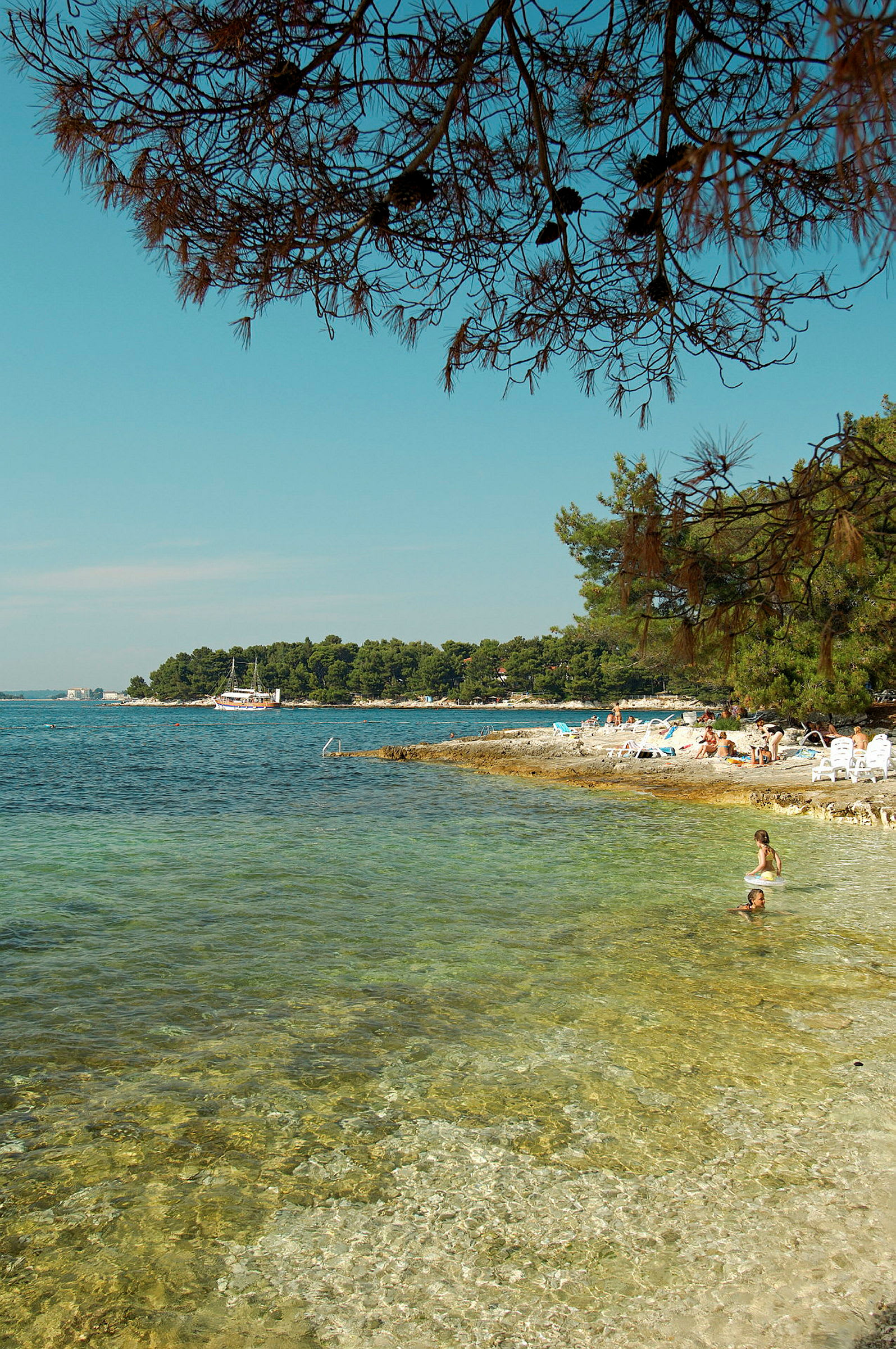 Strand med klart vand og mennesker, der bader, omgivet af træer og en båd længere væk. (AI-genereret tekst)