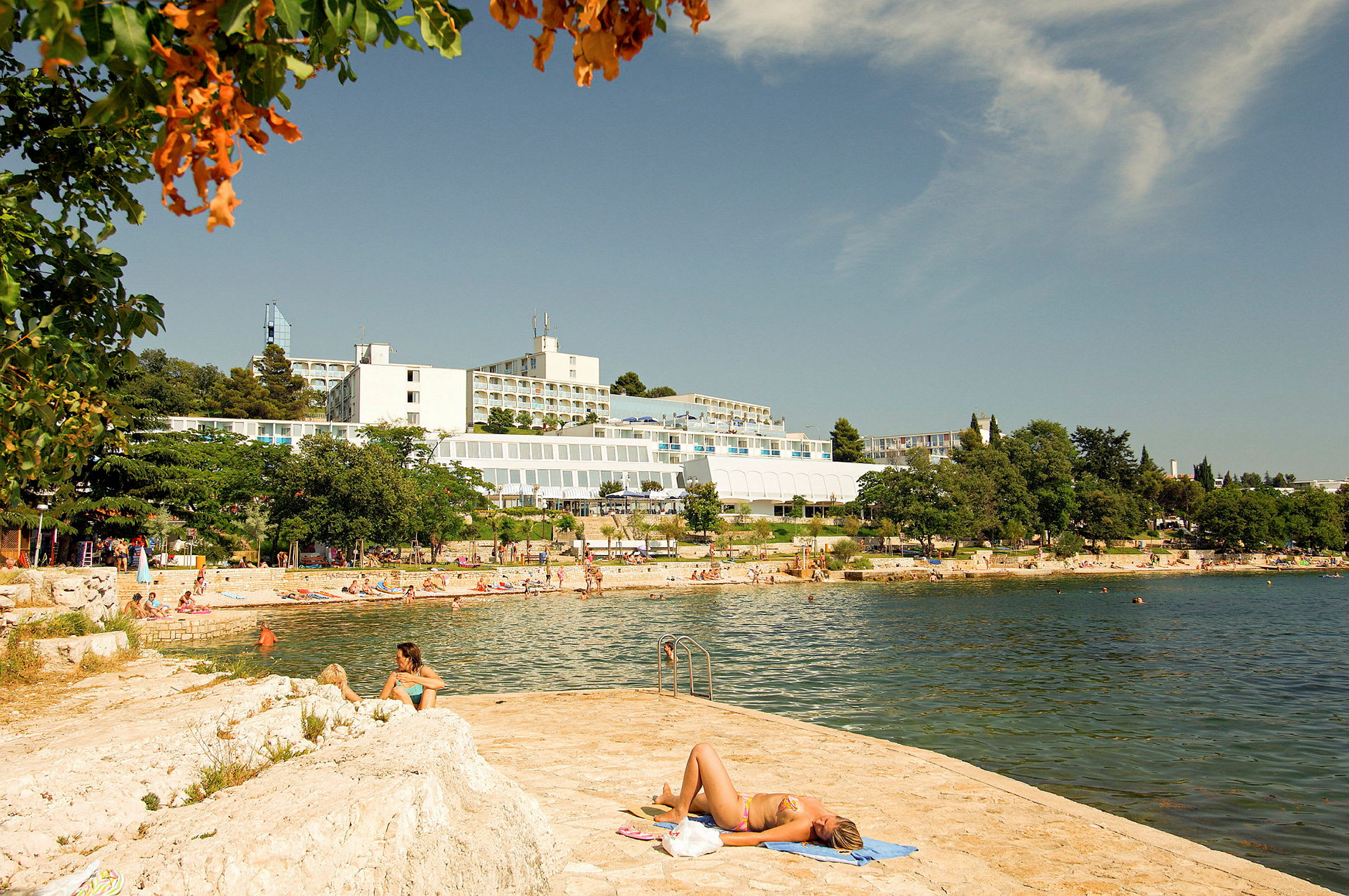 En person ligger på et håndklæde ved vandet med hotelbygninger og en strand i baggrunden. (AI-genereret tekst)