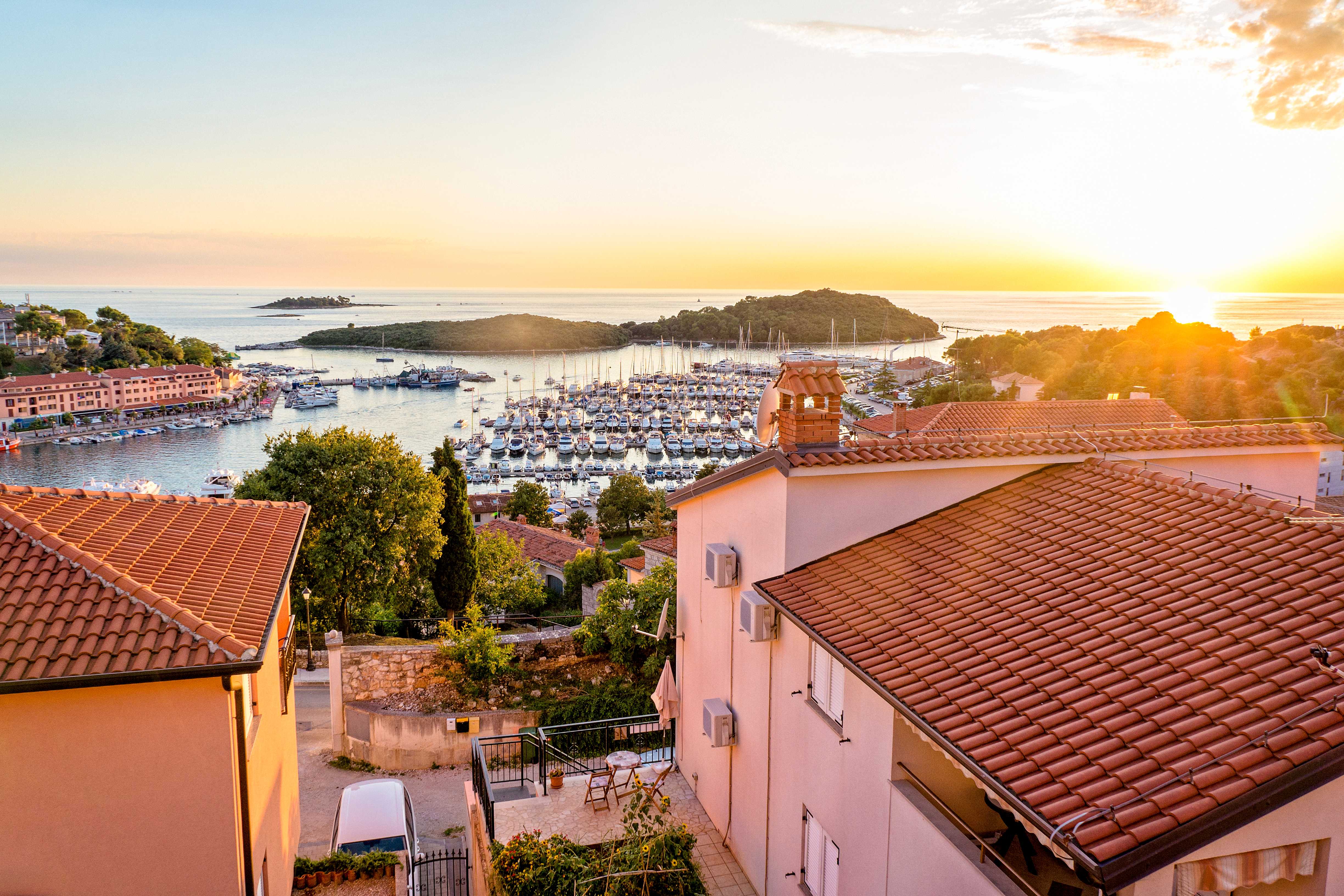 Udsigt over en lystbådehavn med sejlbåde omgivet af huse og grønt ved solnedgang. (AI-genereret tekst)