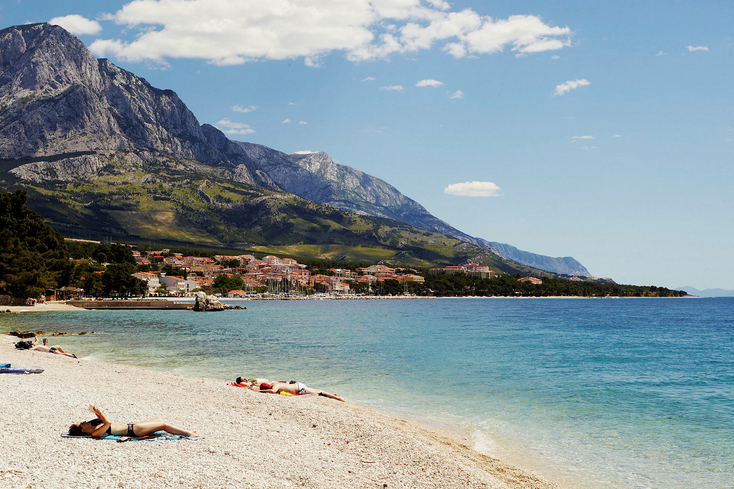 En strandscene med personer, der soler sig, kystby i baggrunden og bjerge. (AI-genereret tekst)