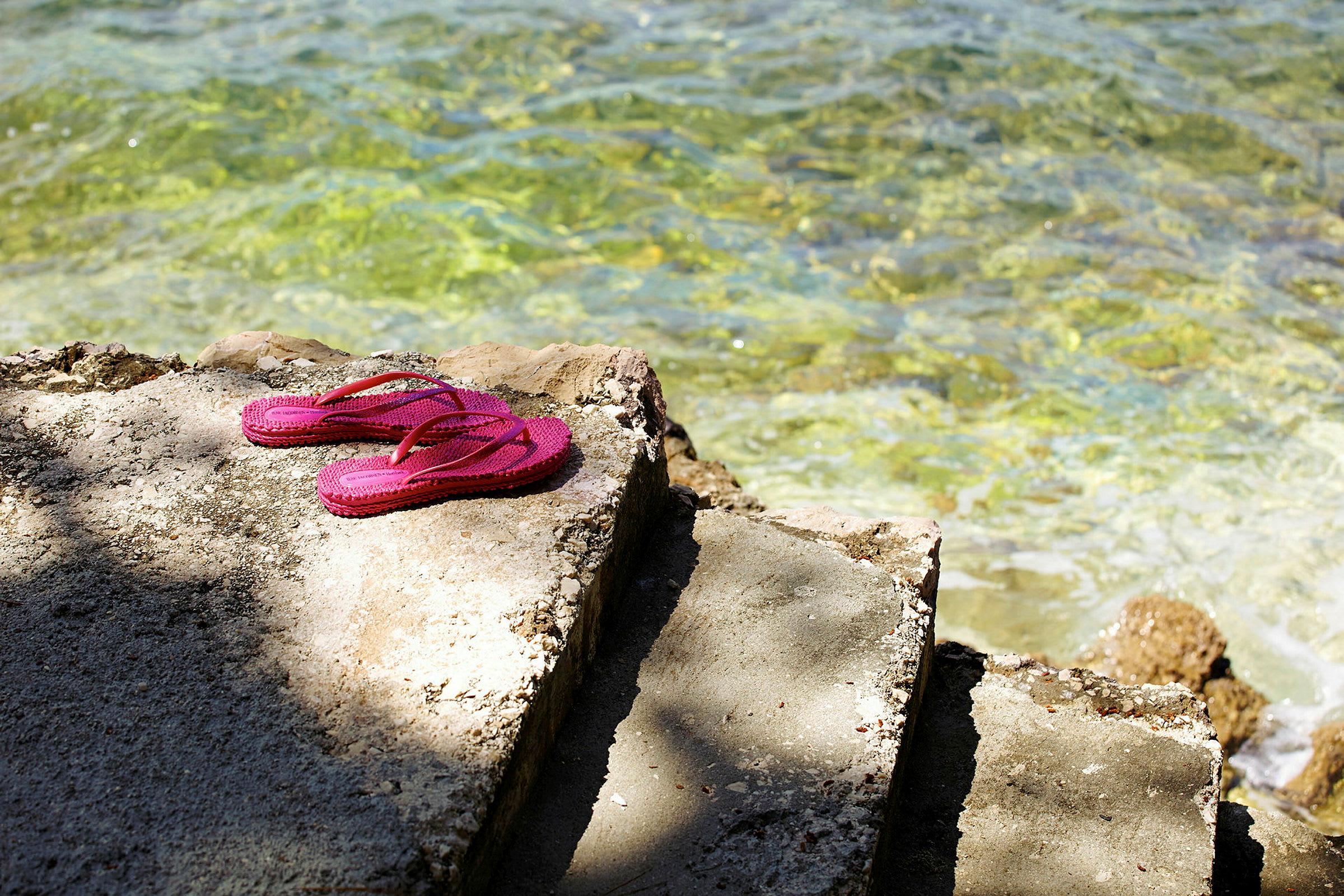 Rosa flip-flops ligger på en cementtrappe ved klart vand. (AI-genereret tekst)