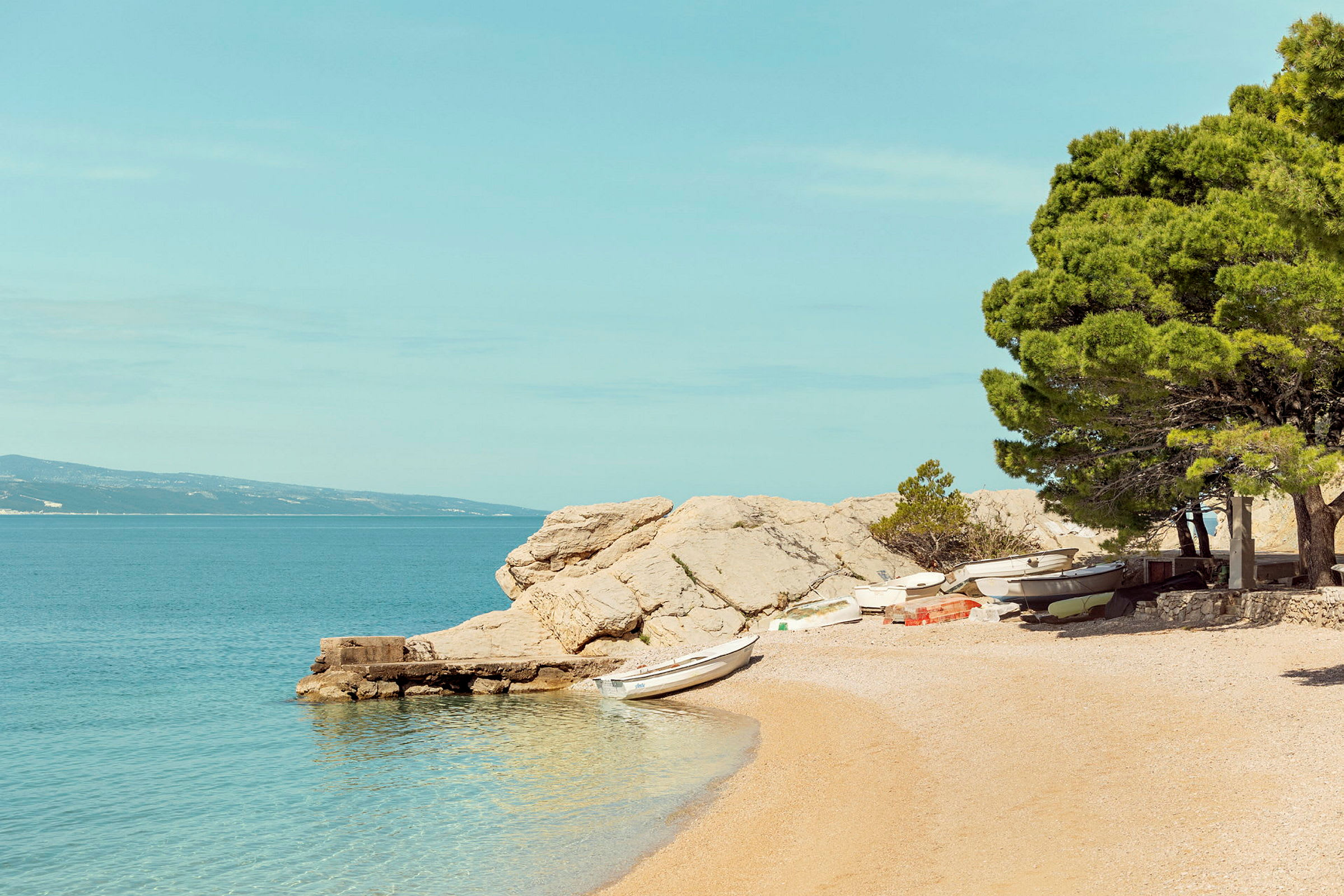 En strand med småbåde, træer og klipper ved vandet. (AI-genereret tekst)