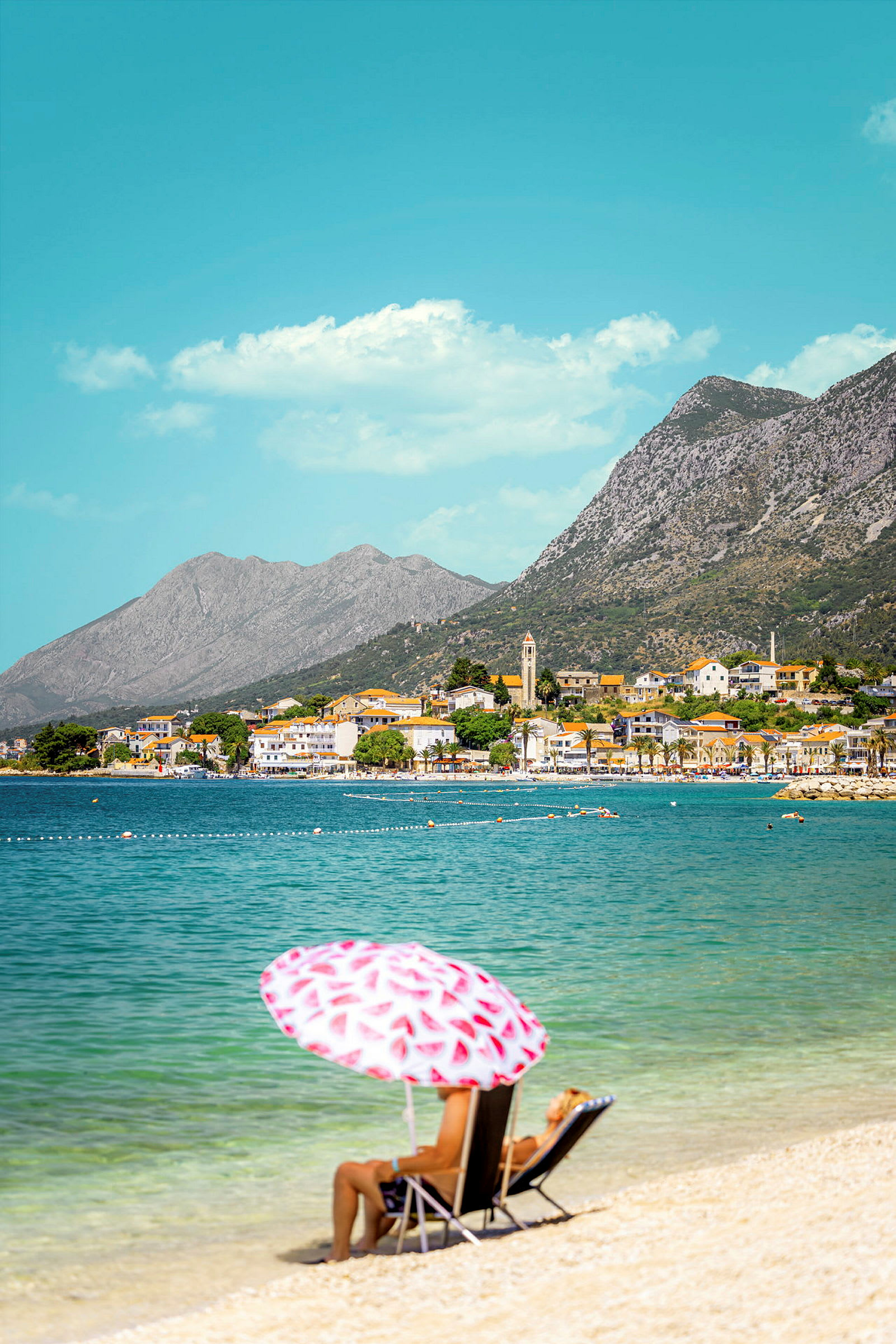 En kvinde i en liggestol under en parasol på en strand med klart vand, omgivet af bjerge og en kystby i baggrunden. (AI-genereret tekst)