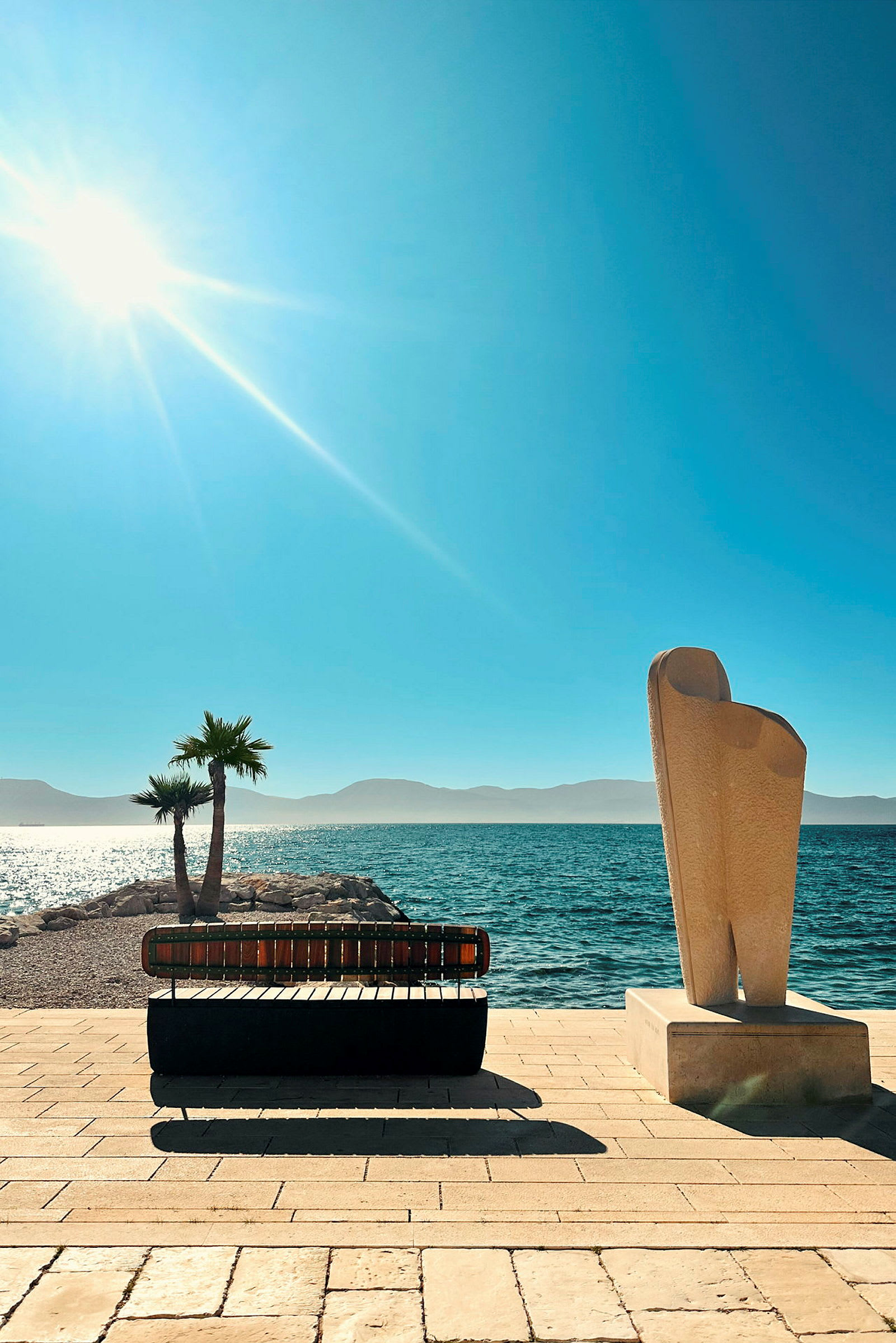 En strandpromenade med en solbænk og en skulptur, med havet og solen i baggrunden. (AI-genereret tekst)