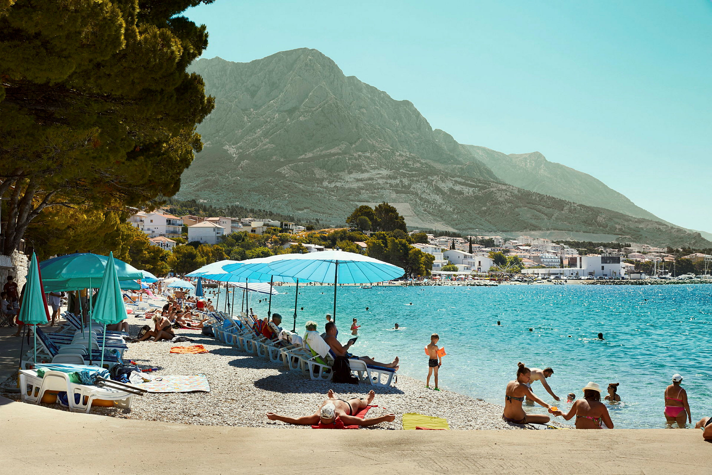 Strand med parasoller og liggestole ved klart vand, omgivet af bjerge. (AI-genereret tekst)