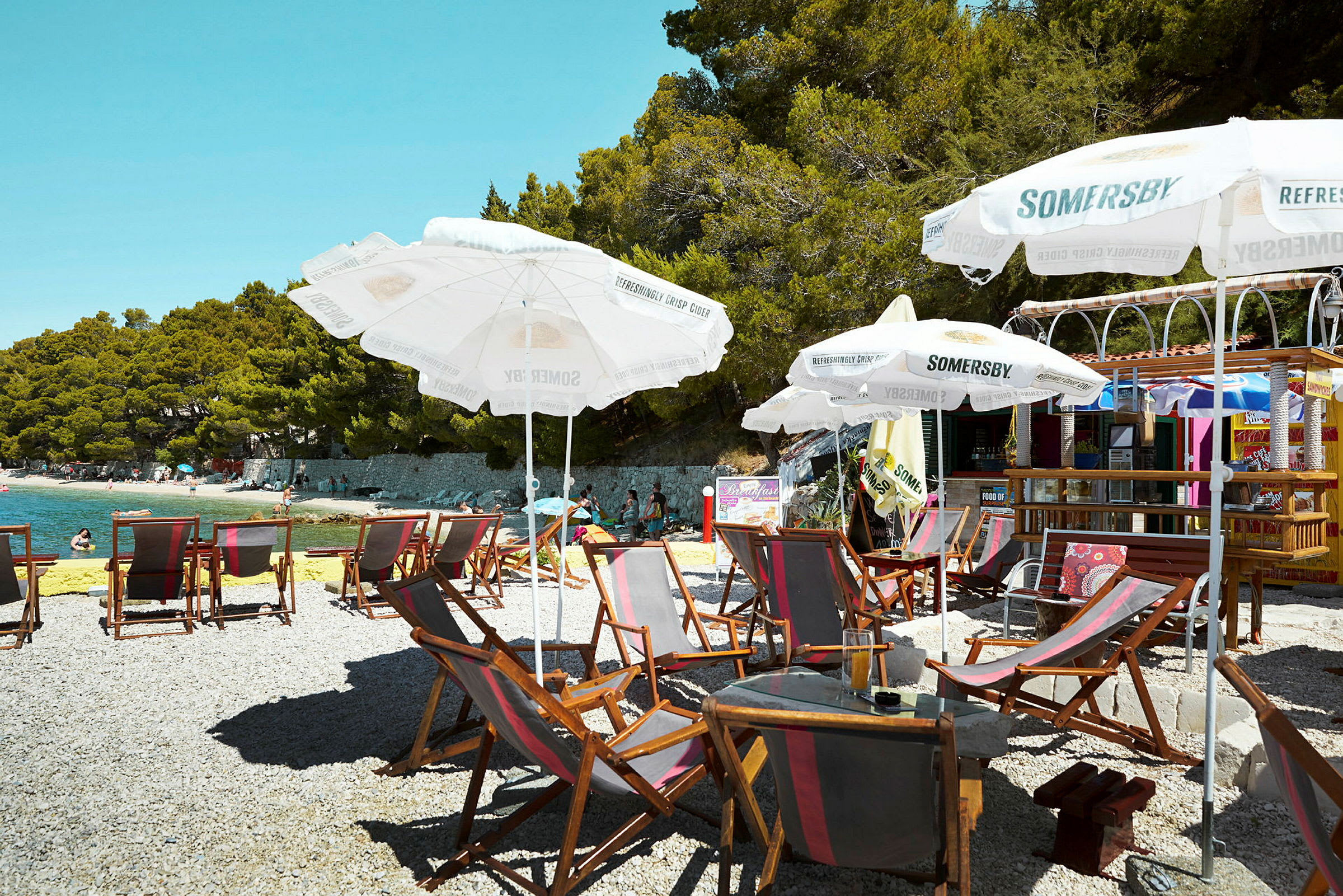 Strand med liggestole, parasoller og en café i baggrunden. (AI-genereret tekst)