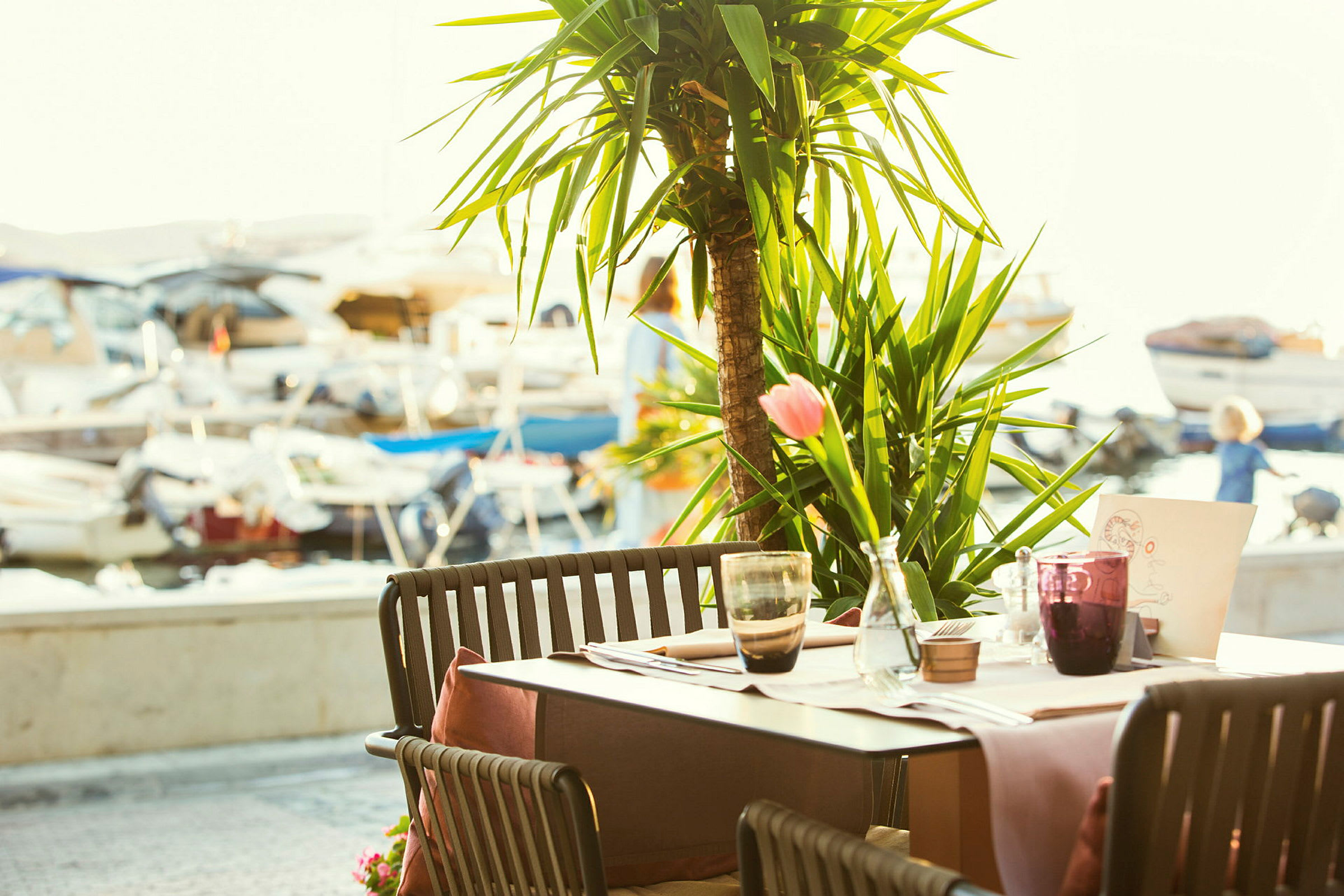 En udendørs restaurant med borde og stole, med en palme i baggrunden og udsigt til små både ved en kaj. (AI-genereret tekst)
