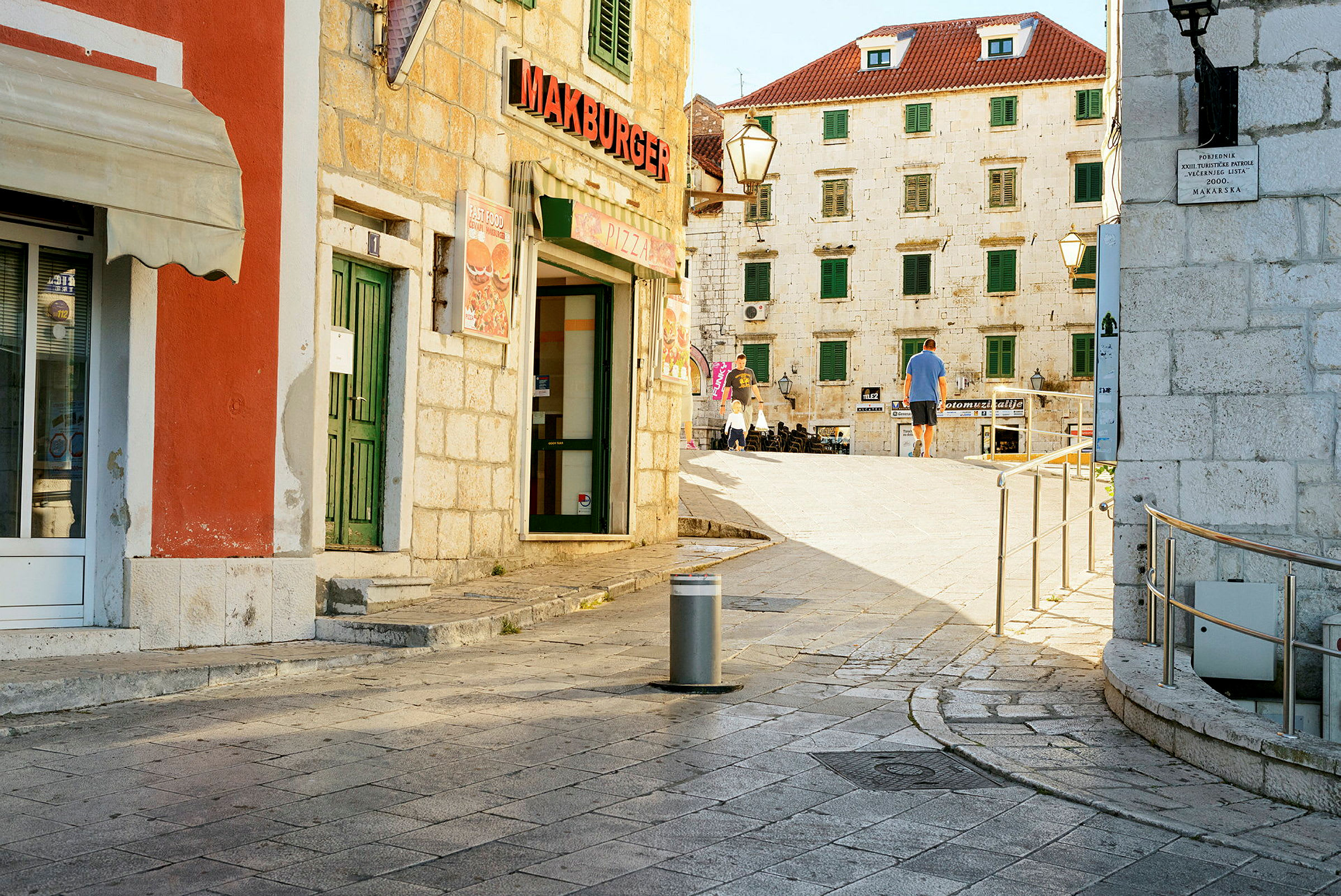 Bymiljø med stenbelagt gade og bygninger med røde tage og grønne skodder samt en restaurantskilt med teksten 'Makburger'. (AI-genereret tekst)