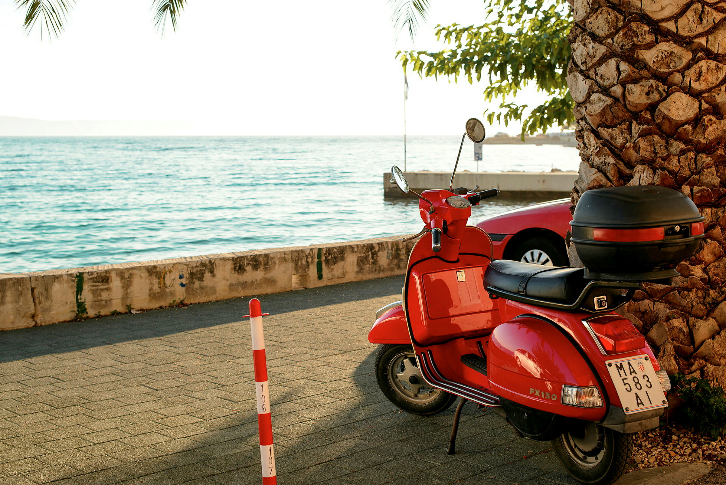 Rød scooter parkeret på en gangsti tæt på havet, omgivet af en palme og parkerede biler. (AI-genereret tekst)